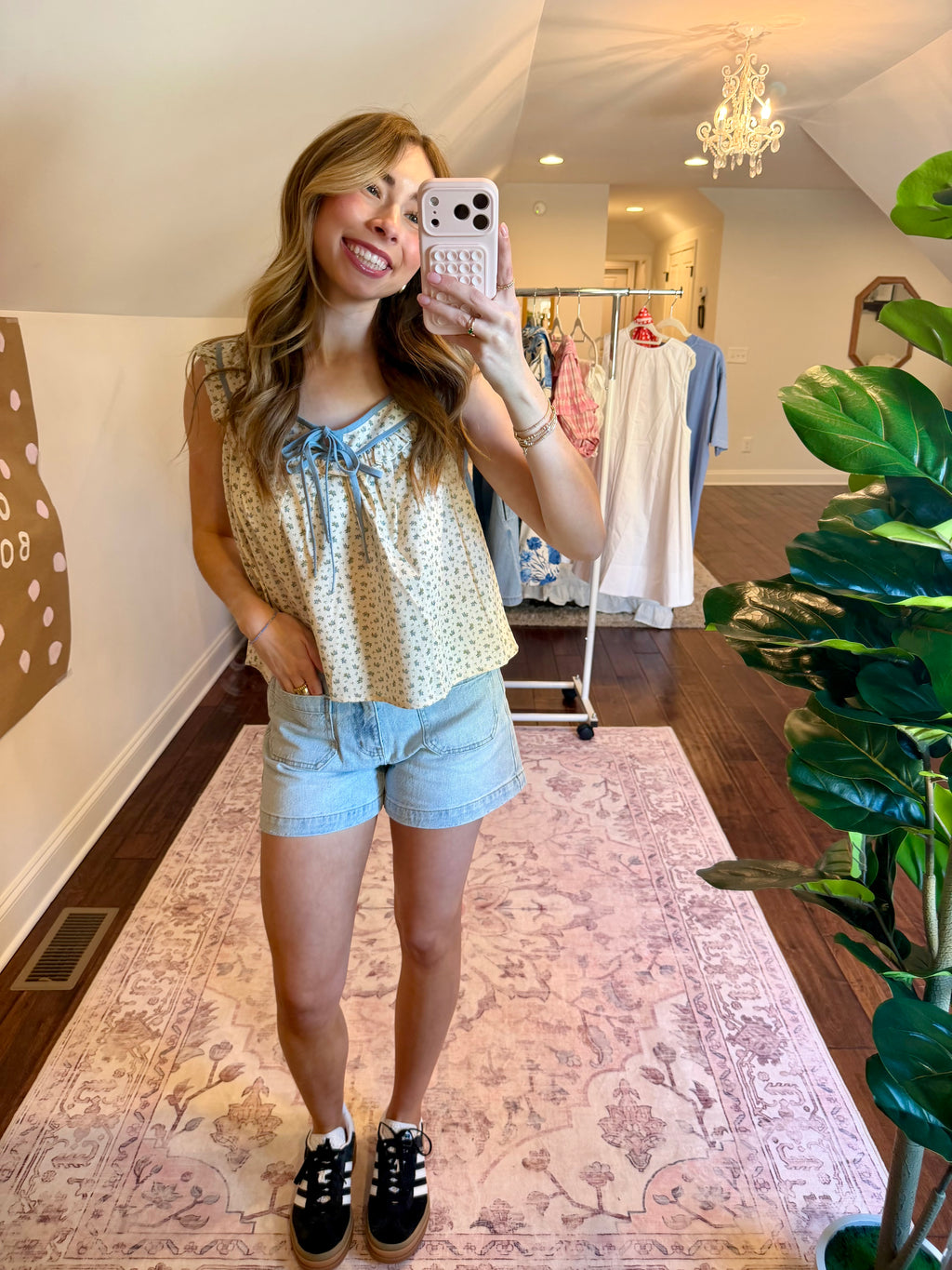 Woman taking a mirror selfie wearing a floral top and denim shorts in a clothing store.