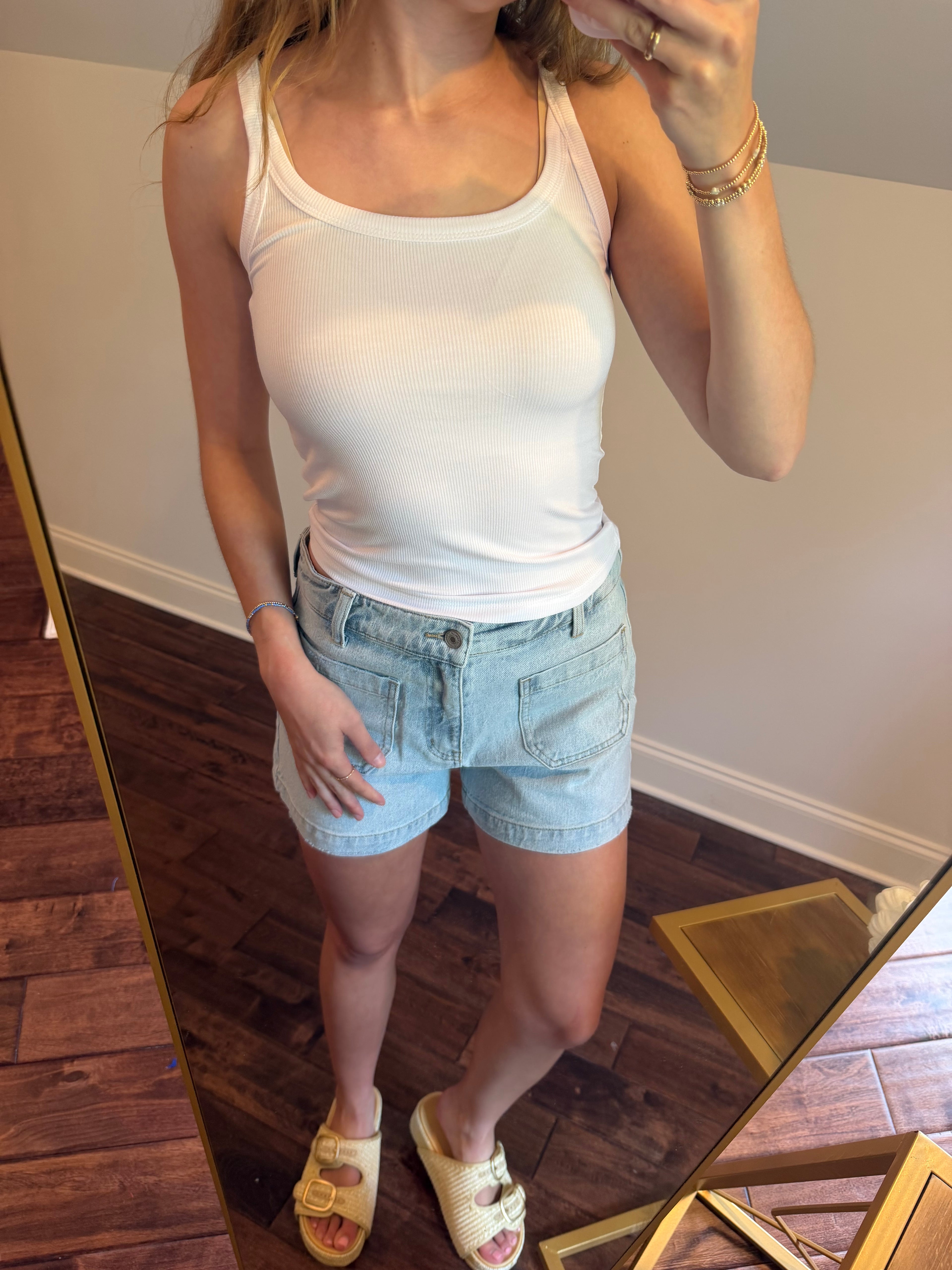 Person wearing a white tank top and light blue denim shorts standing in front of a mirror.