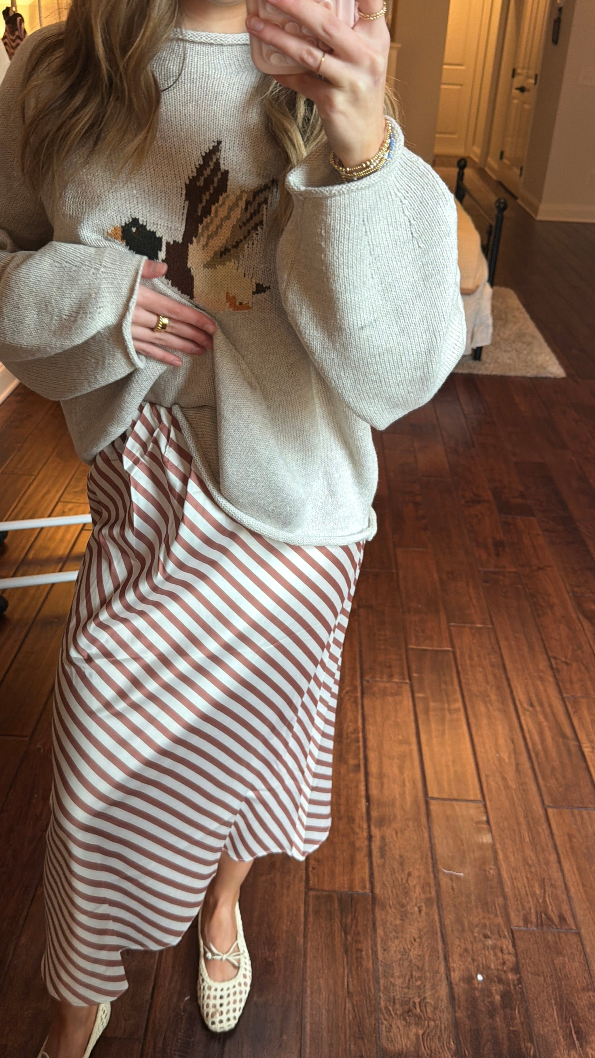 Person wearing a white sweater with a duck design and a brown and white striped skirt indoors.