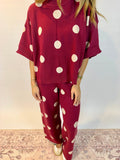 A woman wearing a matching burgundy knit two-piece set with large white polka dots, featuring a short-sleeve top and wide-leg pants, standing against a plain light wall.
