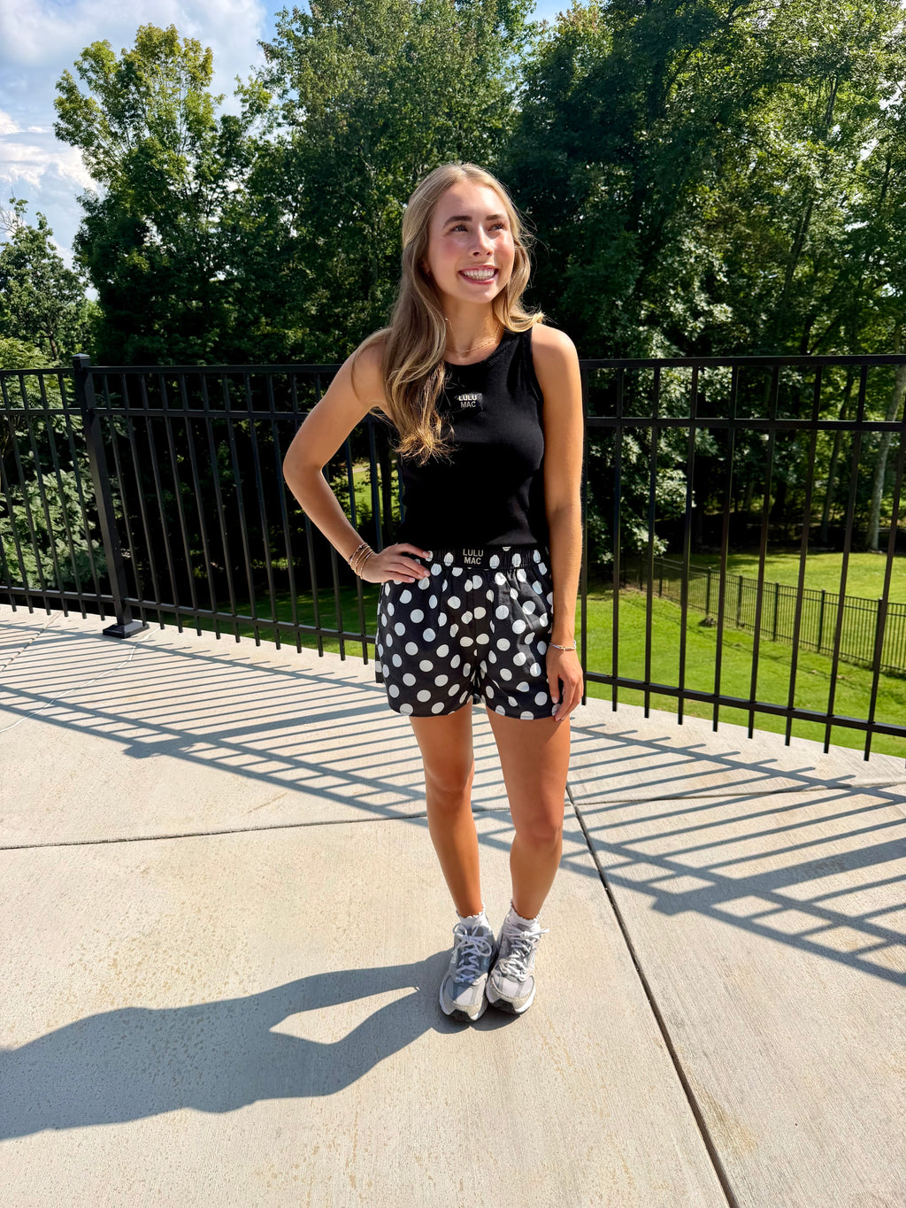 A woman stands outdoors on a concrete walkway wearing a black sleeveless top and black shorts with white polka dots, paired with gray sneakers. Trees and a black metal railing are visible in the background.