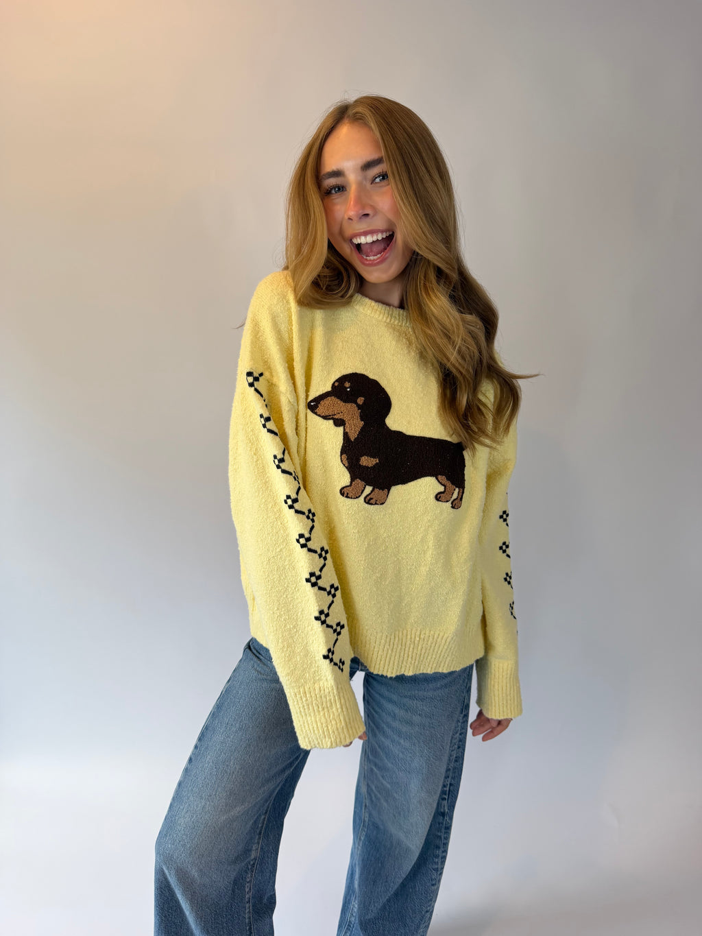 A woman modeling a pale yellow knit sweater featuring a black and tan dachshund graphic on the front, with black geometric patterns along the sleeves, paired with blue jeans against a plain light gray background.