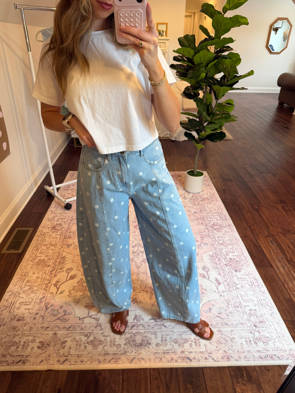 Woman taking a mirror selfie wearing a white top and light blue pants with a polka dot pattern in a room with a plant and rug.