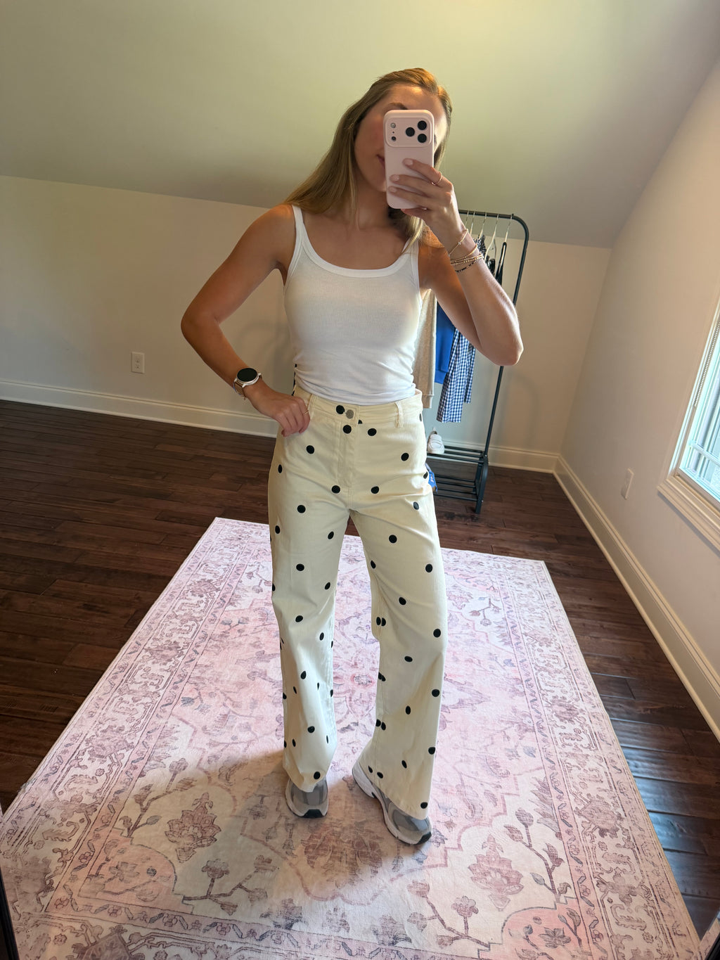 A woman takes a mirror selfie in a room, wearing a white ribbed tank top and cream-colored wide-leg pants with black polka dots. She is standing on a pink patterned area rug with dark wood floors and a clothing rack in the background.
