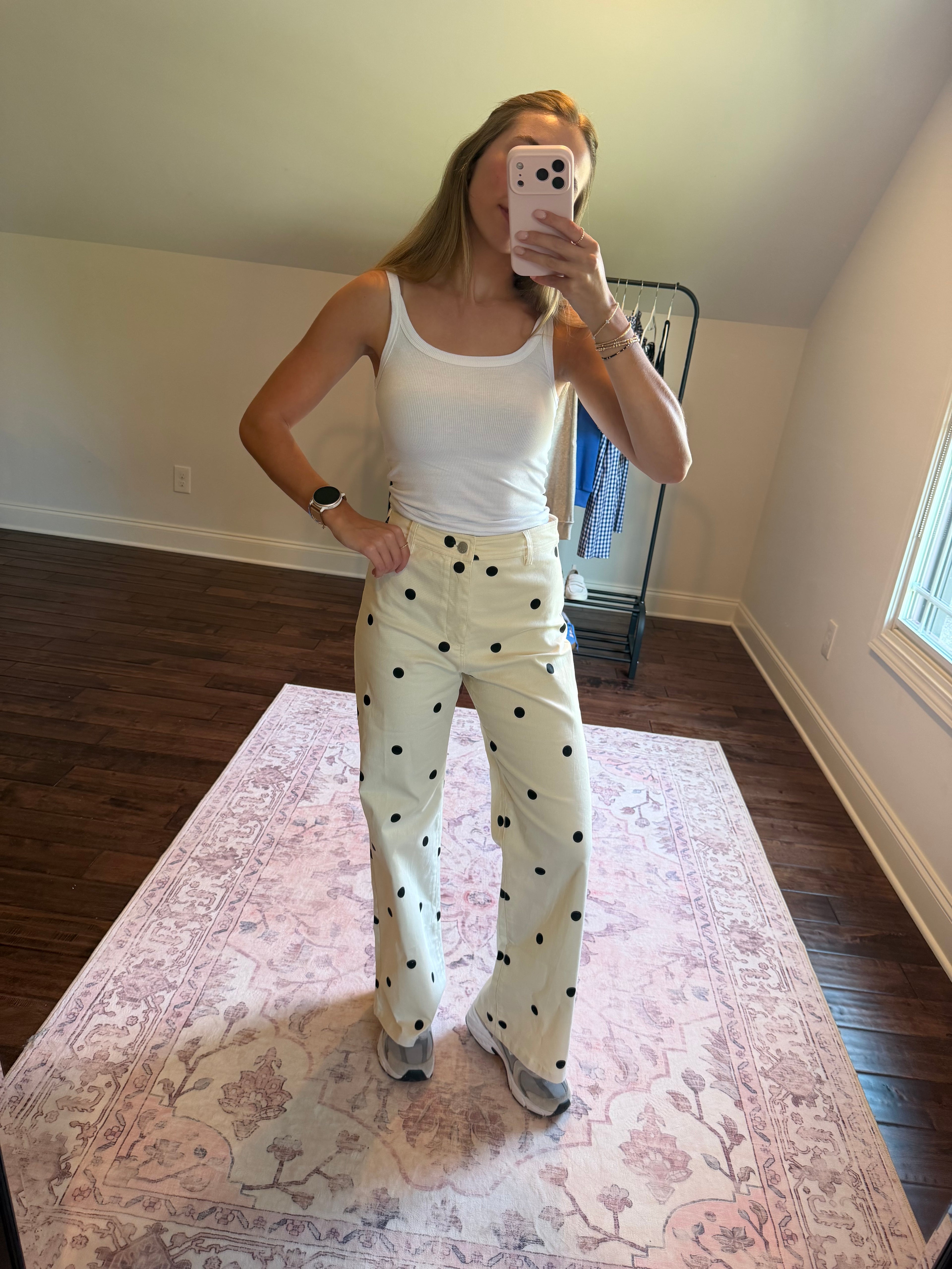 A woman takes a mirror selfie in a room, wearing a white ribbed tank top and cream-colored wide-leg pants with black polka dots. She is standing on a pink patterned area rug with dark wood floors and a clothing rack in the background.
