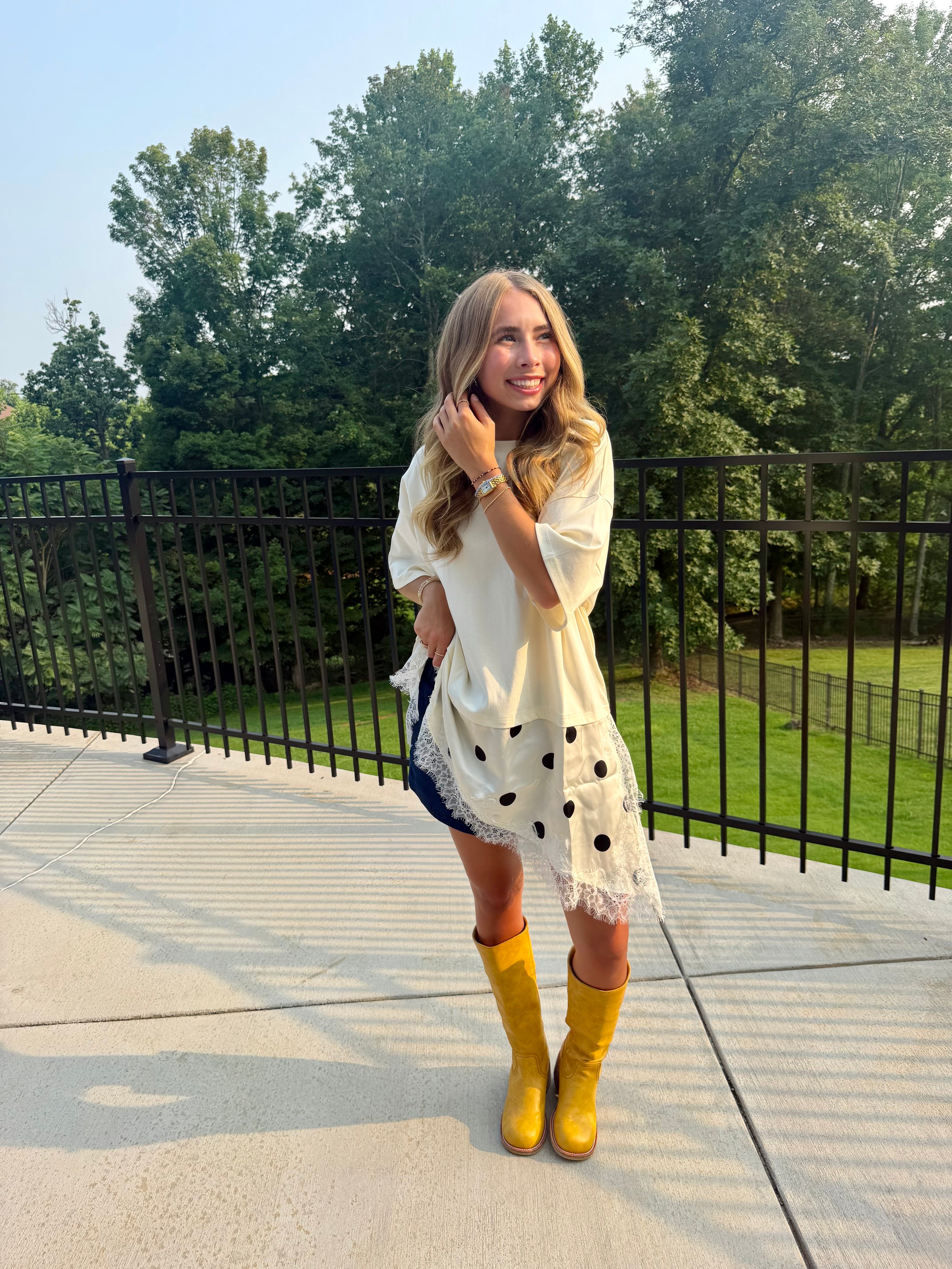 A woman stands outdoors on a concrete patio wearing a white long-sleeve tunic top with black polka dots and fringe, a dark denim skirt, and bright yellow rain boots, with a black metal railing and green trees in the background.