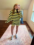 A woman takes a mirror selfie in a room wearing a yellow and black striped oversized sweater and matching shorts. She is standing on a pink patterned area rug with hardwood floors, a window, and a clothing rack in the background.