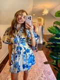 Woman taking a mirror selfie wearing a blue floral dress in a room with plants and a chandelier.