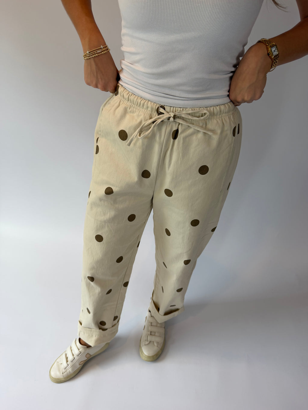 A person wearing beige drawstring pants with dark brown polka dots, paired with a white ribbed tank top and white sneakers, standing against a light gray studio background.