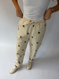 A person wearing beige drawstring pants with dark brown polka dots, paired with a white ribbed tank top and white sneakers, standing against a light gray studio background.