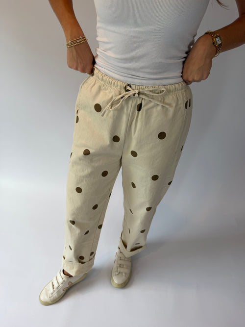 A person wearing beige drawstring pants with dark brown polka dots, paired with a white ribbed tank top and white sneakers, standing against a light gray studio background.