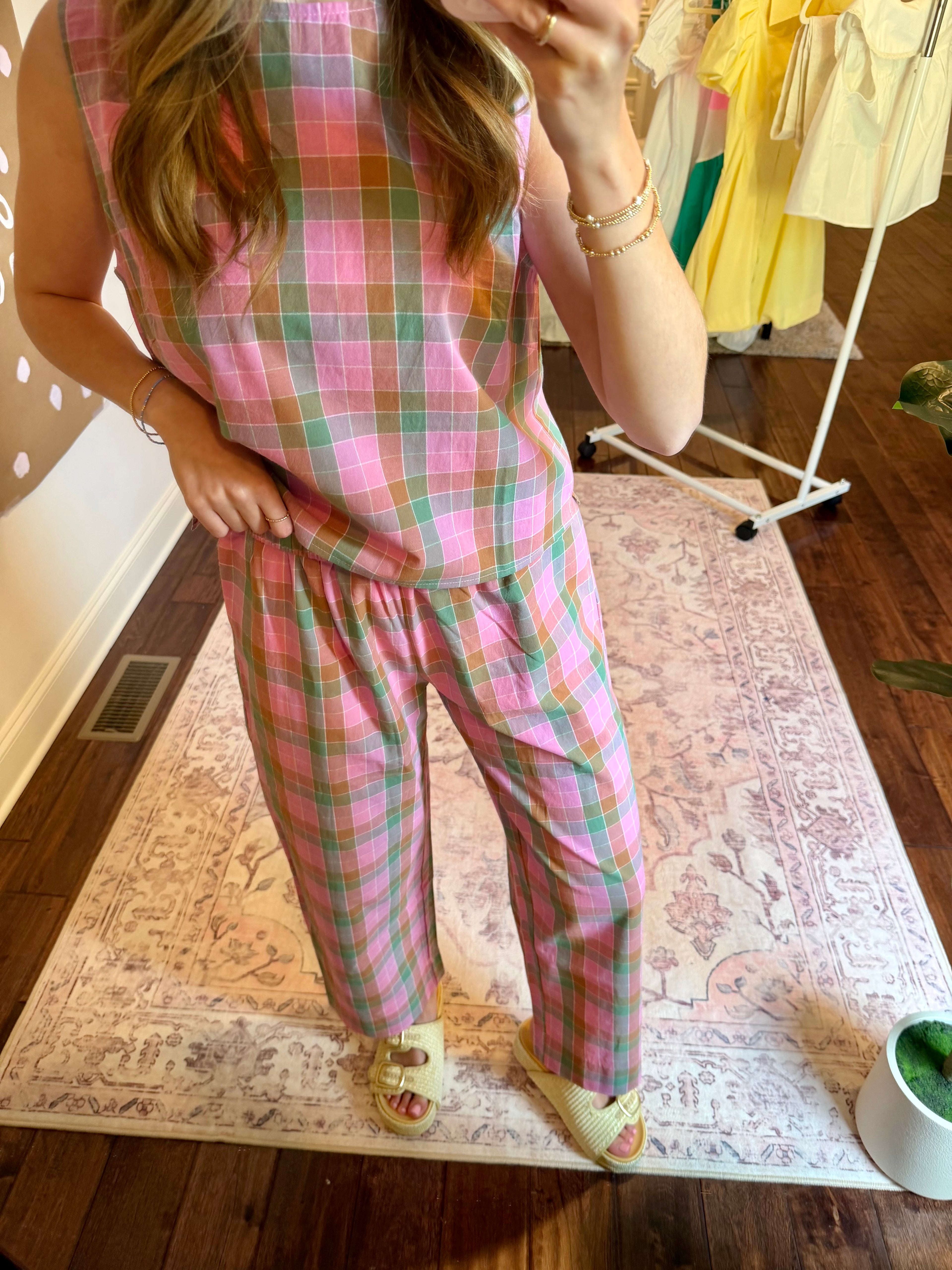 Person wearing a pink plaid outfit taking a mirror selfie in a room with a clothing rack in the background.