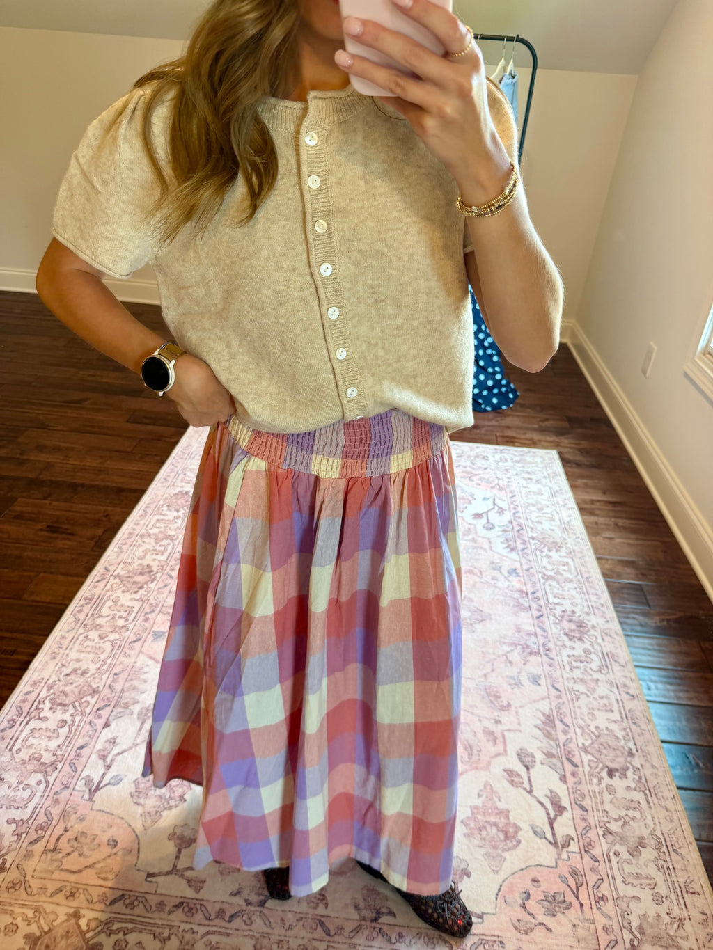A woman takes a mirror selfie in a room, wearing a beige short-sleeve button-front knit top and a pink, purple, and white plaid midi skirt.