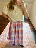 A woman takes a mirror selfie in a room, wearing a beige short-sleeve button-front knit top and a pink, purple, and white plaid midi skirt.