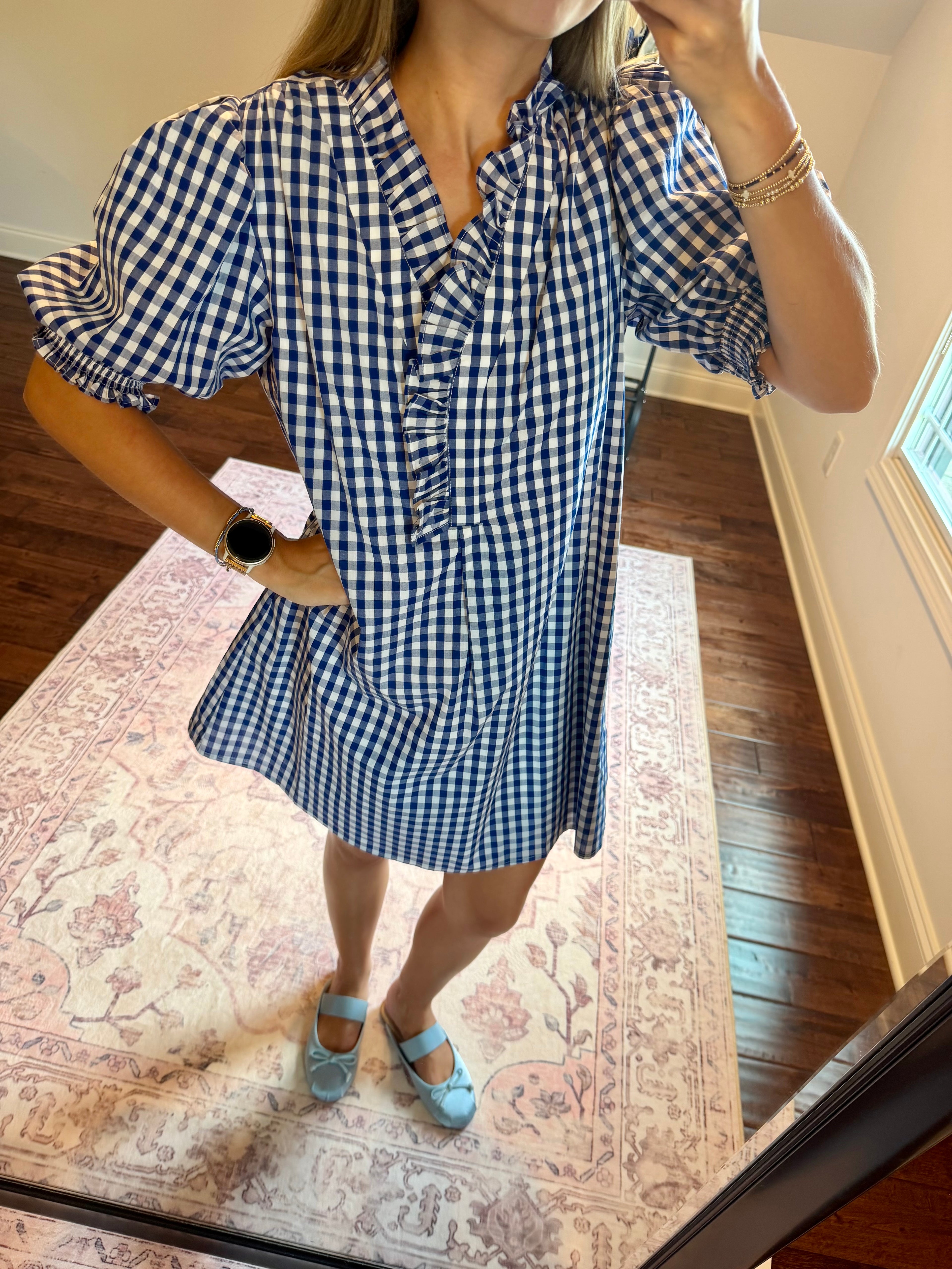 A woman takes a mirror selfie wearing a blue and white gingham mini dress with puff sleeves and a ruffled collar. She is standing on a patterned rug in a room with hardwood floors.