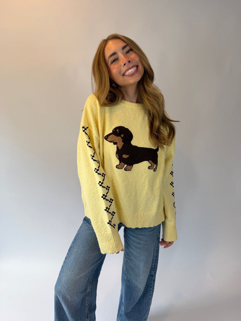 A woman wearing a pale yellow knit sweater with a dachshund graphic and black decorative stripes on the sleeves, paired with blue jeans, posing against a light gray studio background.