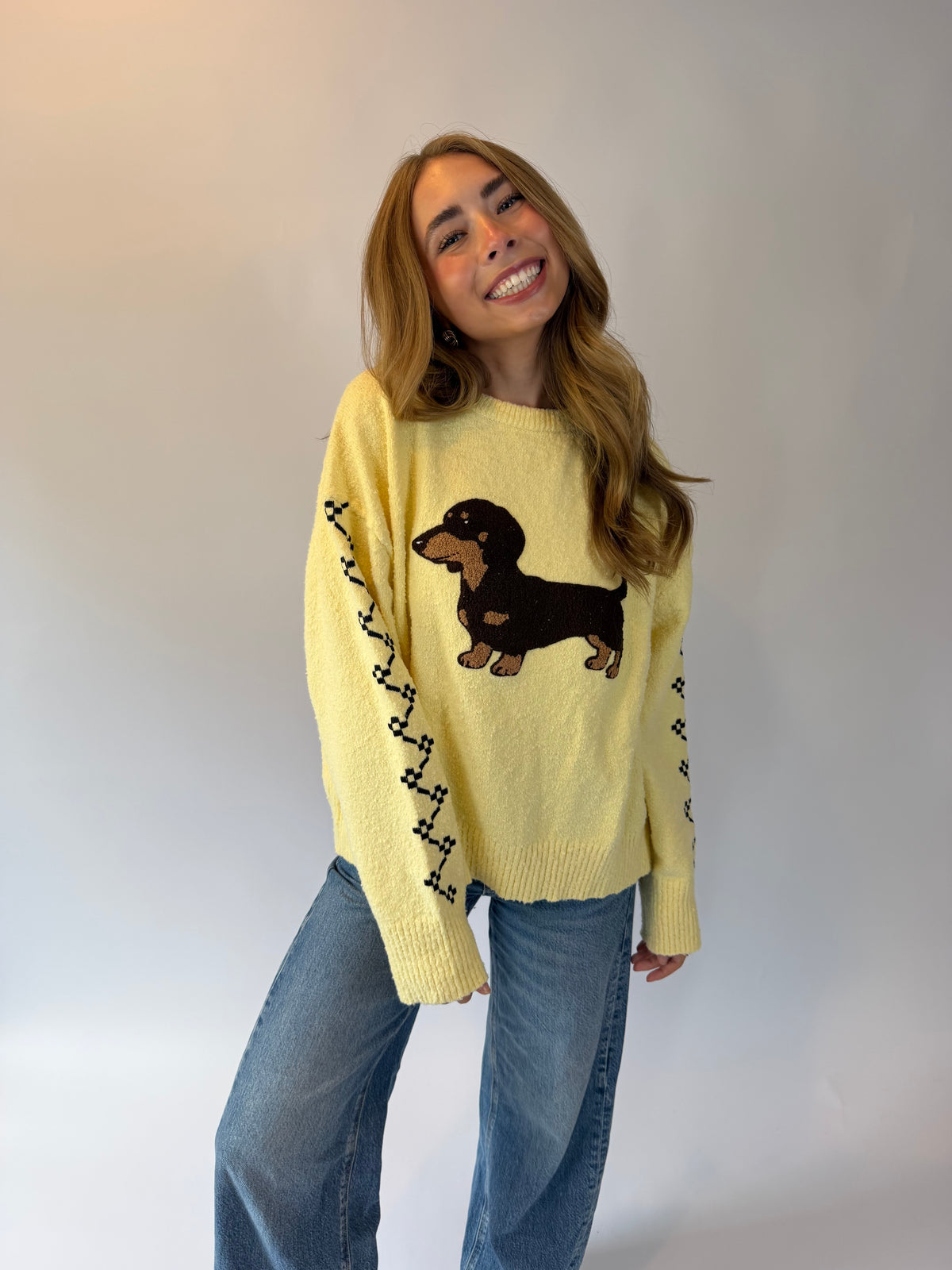 A woman wearing a pale yellow knit sweater with a dachshund graphic and black decorative stripes on the sleeves, paired with blue jeans, posing against a light gray studio background.