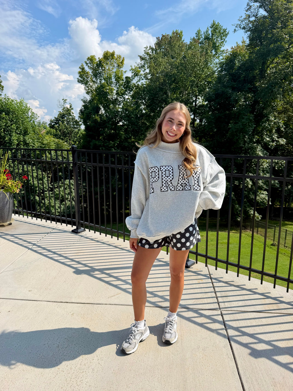 A young woman stands on an outdoor patio wearing a light gray oversized sweatshirt with 'PRAY' in polka dots, black and white polka dot shorts, and white sneakers, with trees and a blue sky in the background.