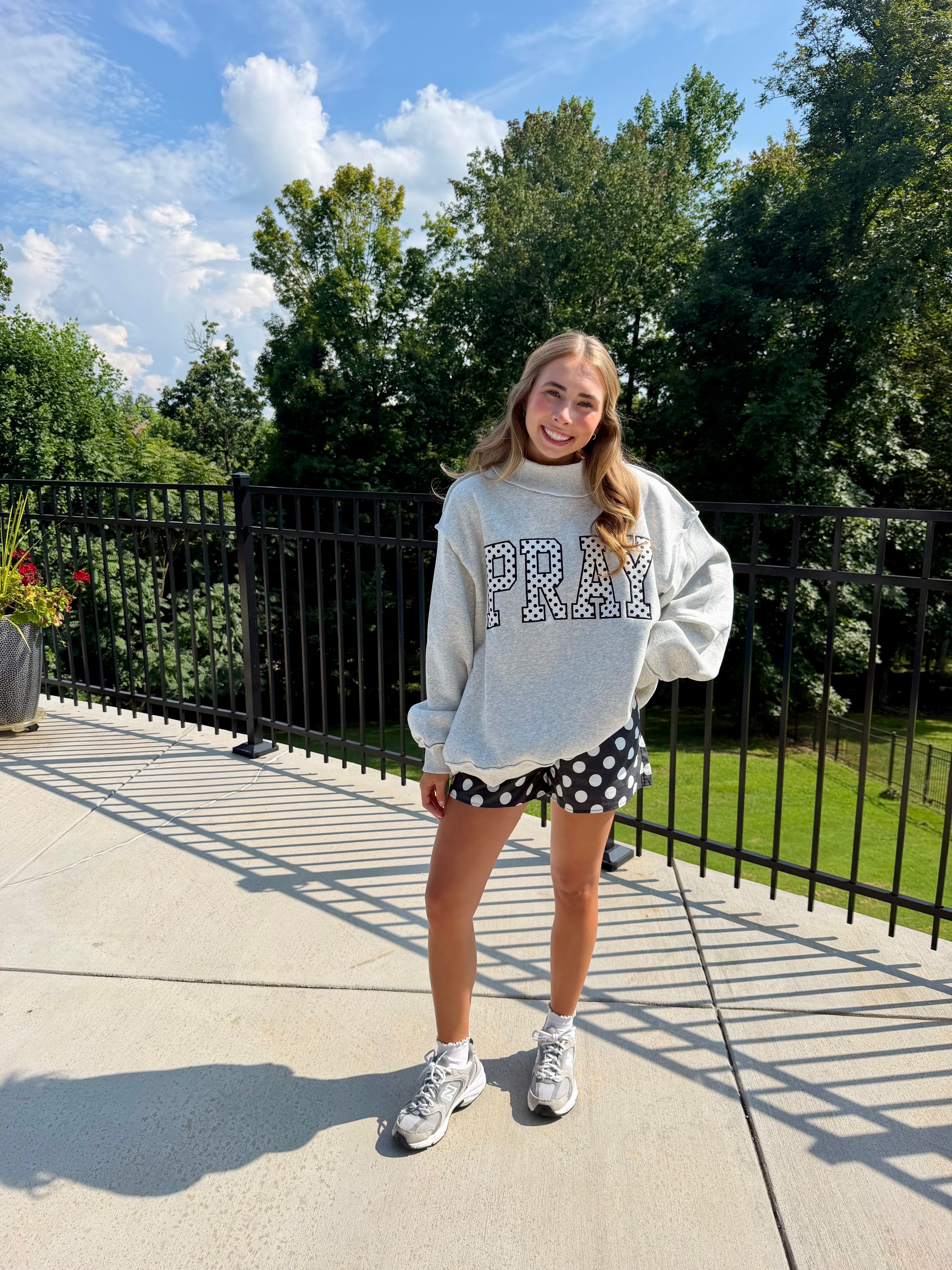 A young woman stands on an outdoor patio wearing a light gray oversized sweatshirt with 'PRAY' in polka dots, black and white polka dot shorts, and white sneakers, with trees and a blue sky in the background.