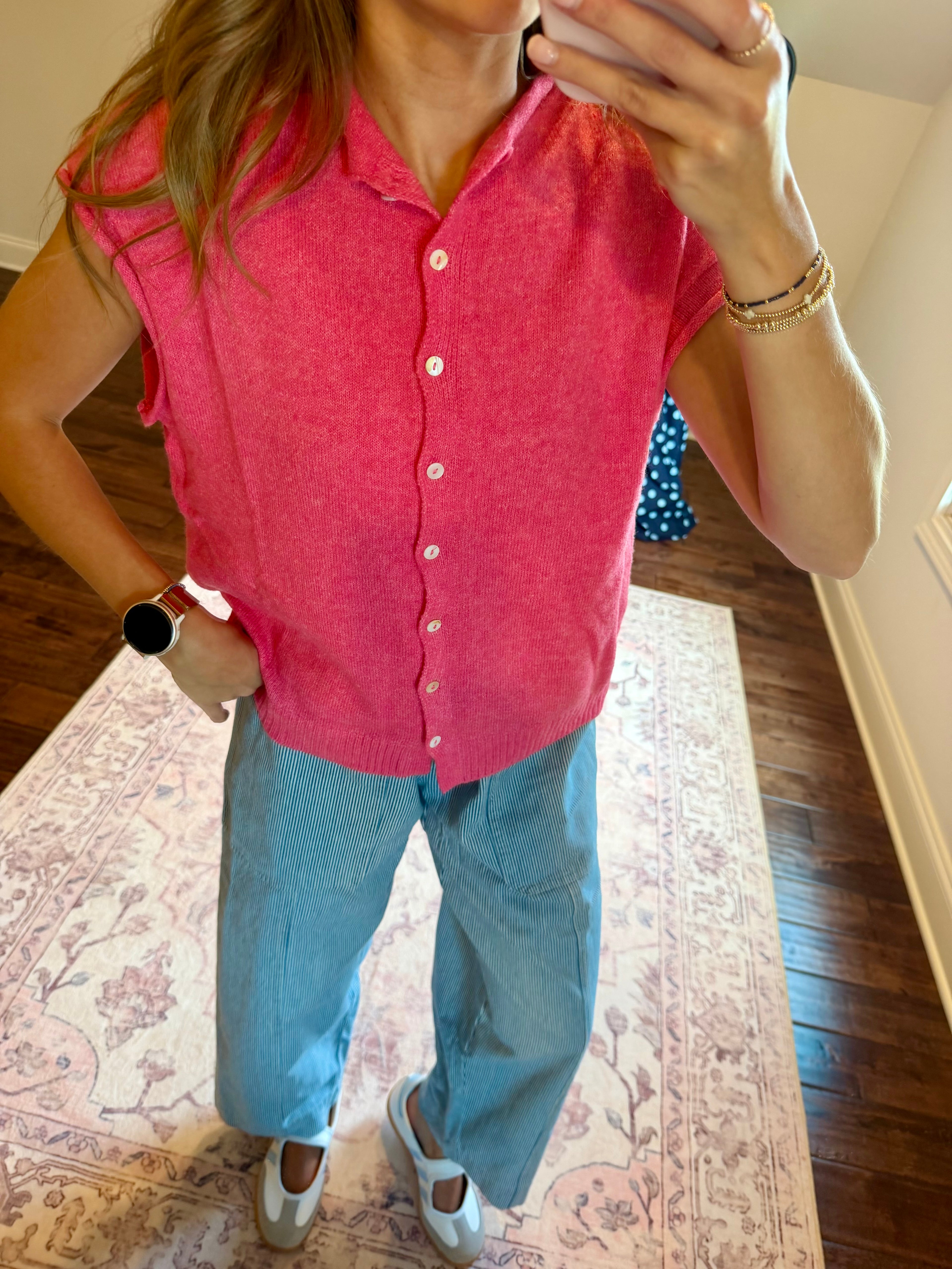 A woman takes a mirror selfie wearing a bright pink sleeveless button-up knit top, light blue wide-leg jeans, and white sneakers in an indoor room with a patterned rug.