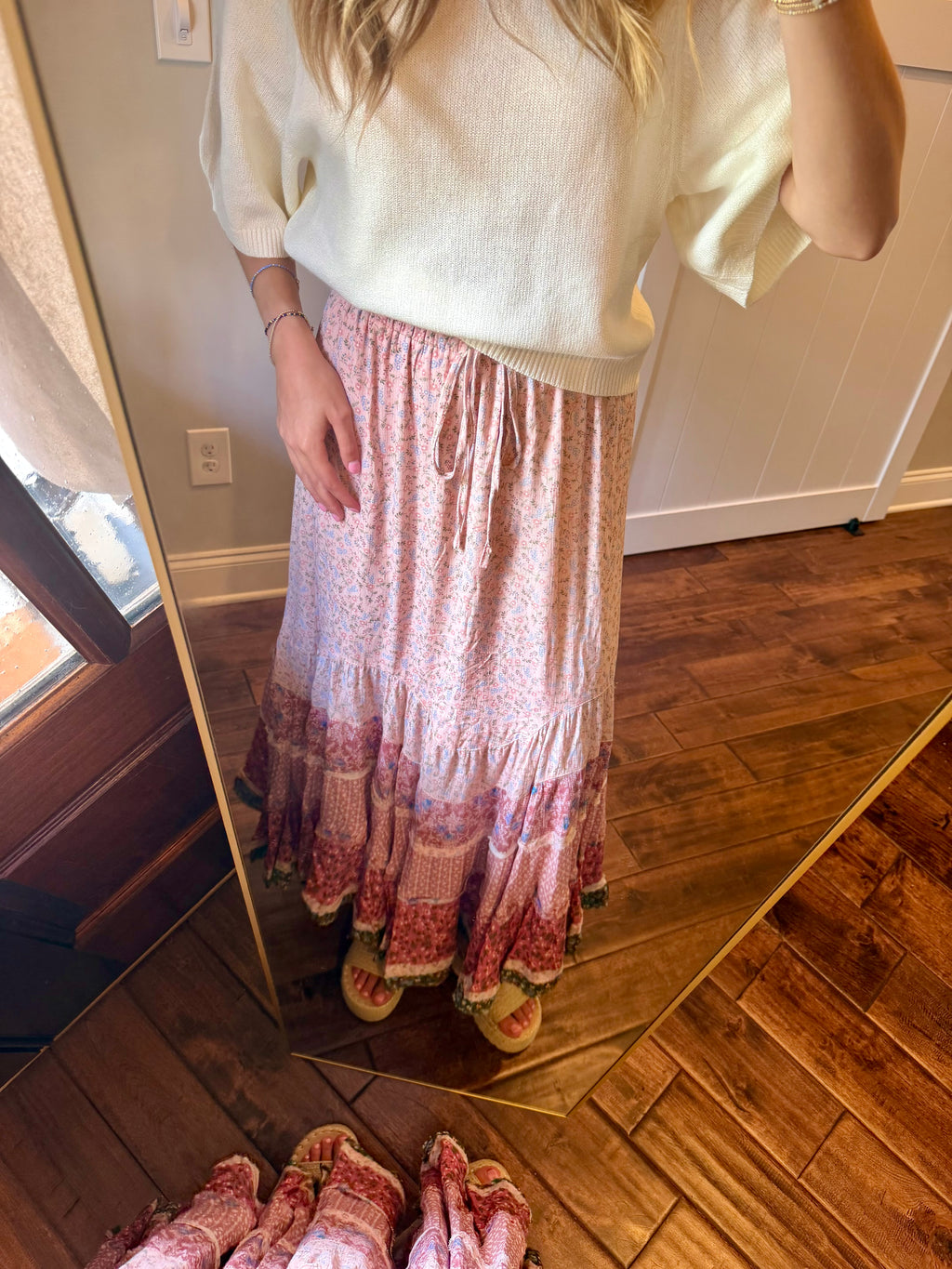 Person wearing a white sweater and patterned skirt standing in front of a mirror on a wooden floor.