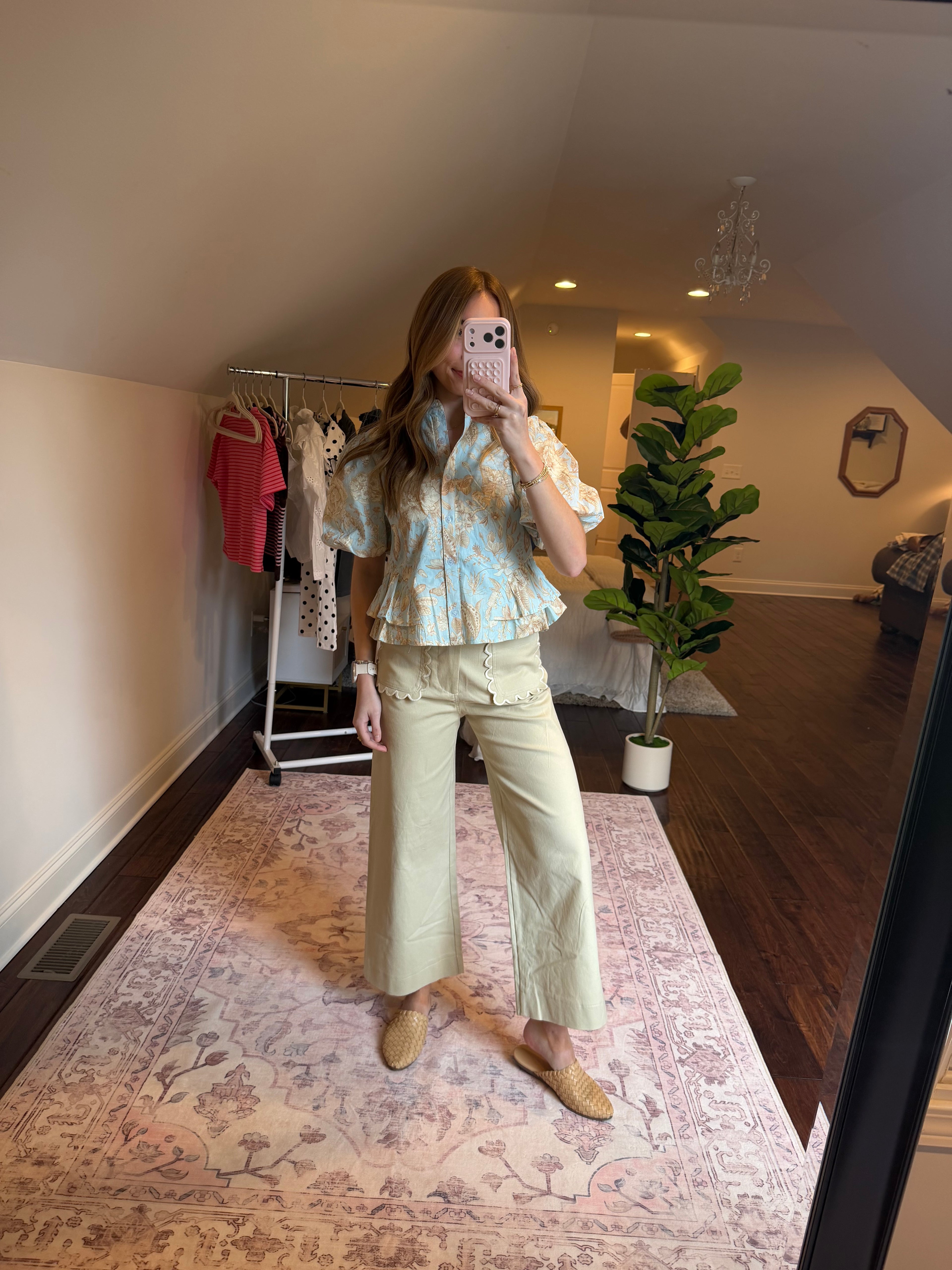 Person wearing a floral top and light-colored pants in a room with patterned wallpaper.