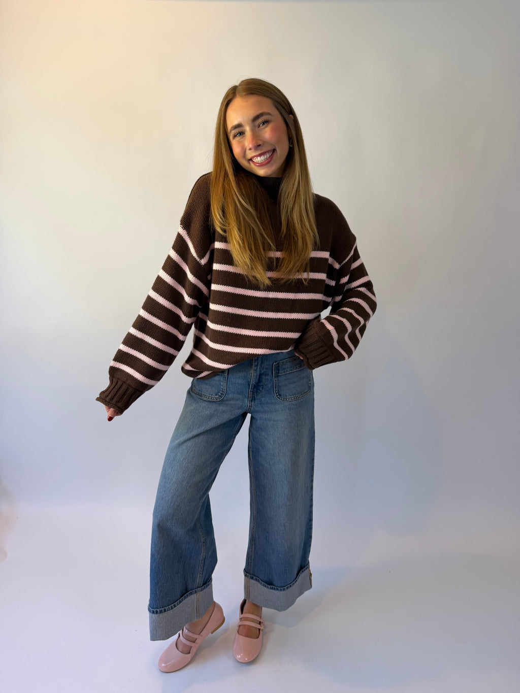 A woman wearing a brown and pink striped knit sweater, blue jeans, and pink ballet flats, photographed against a plain light background.