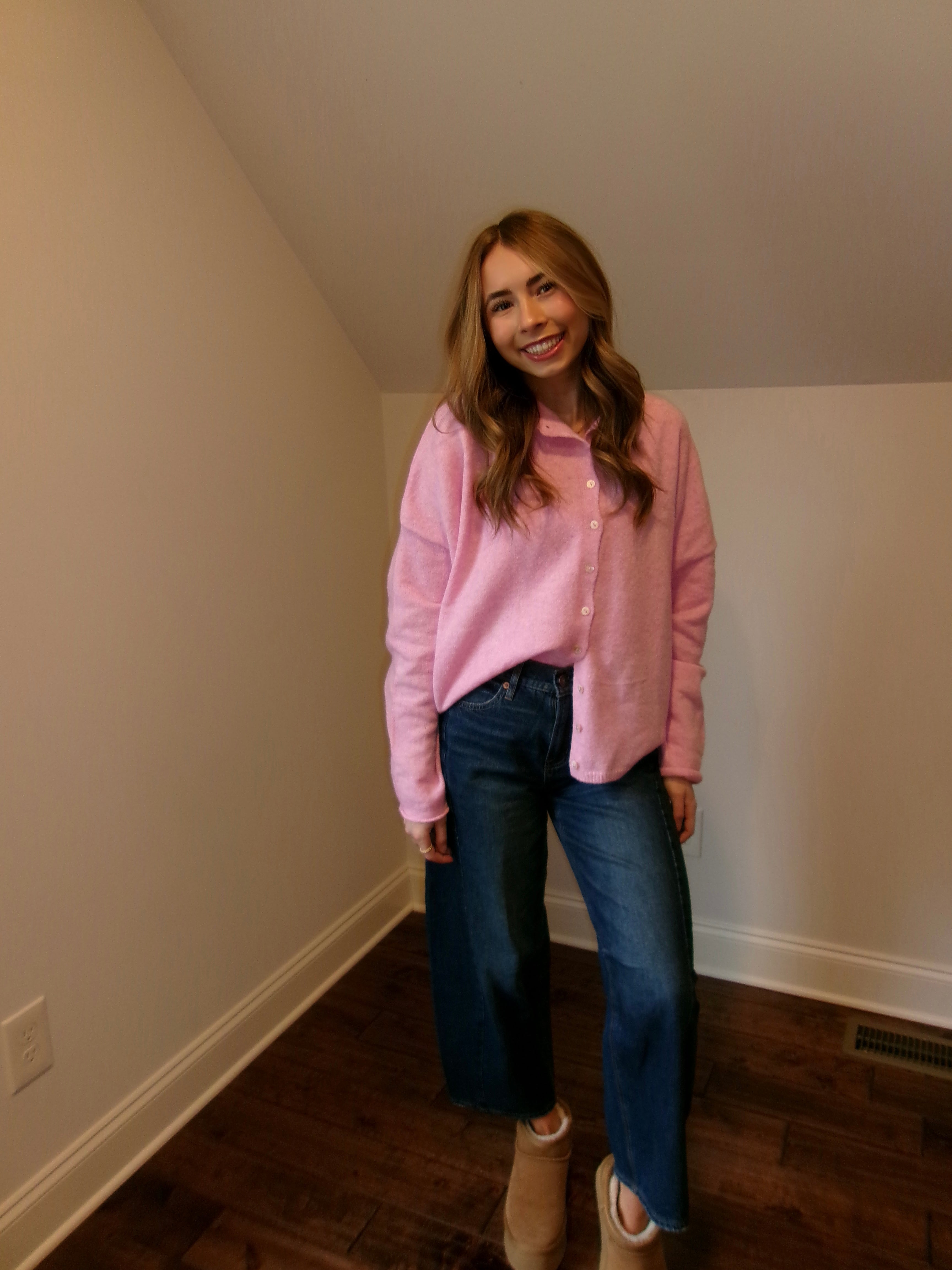 Woman wearing a pink shirt and blue jeans standing in a room with white walls and wooden floor.