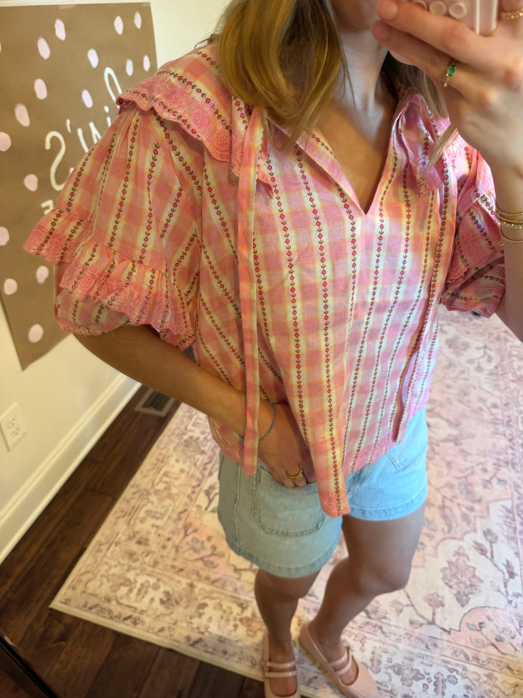 Woman taking a mirror selfie wearing a pink plaid shirt and denim shorts in a room with a chandelier and clothing rack.