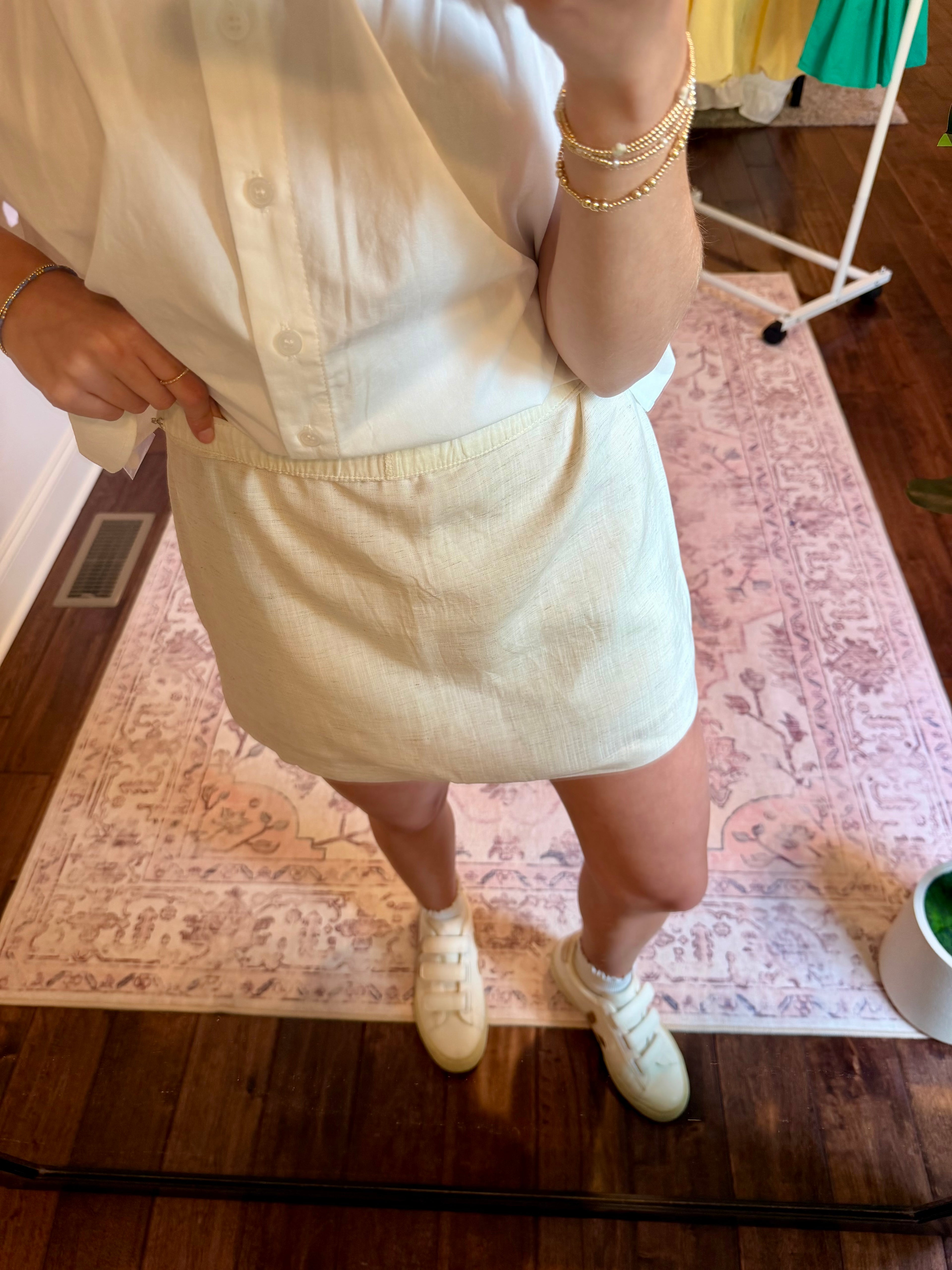 Person wearing a cream skirt and white top with a patterned rug and wooden floor in the background