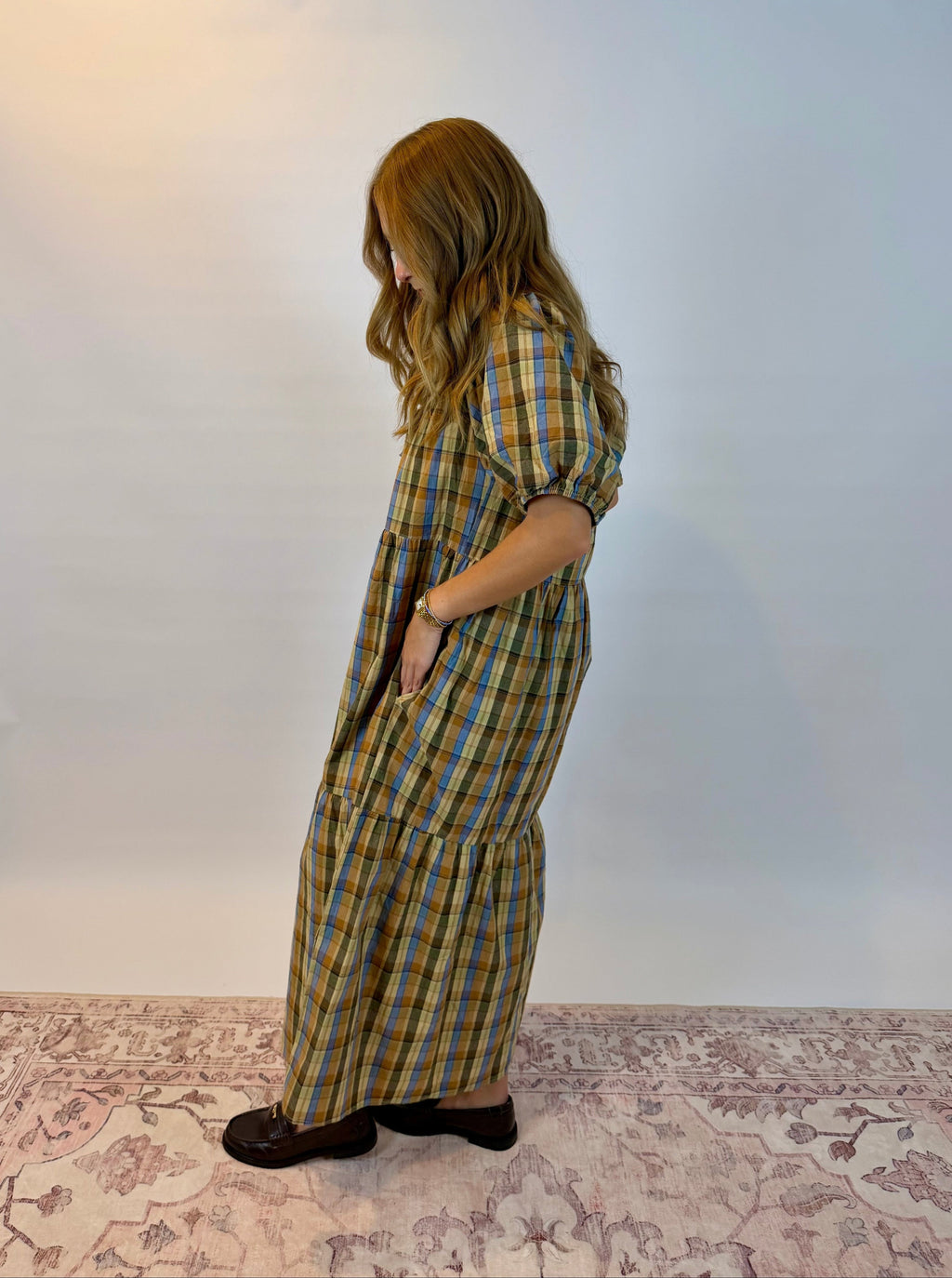 A woman wearing a long-sleeved plaid maxi dress with pockets, standing in profile against a plain white studio background.