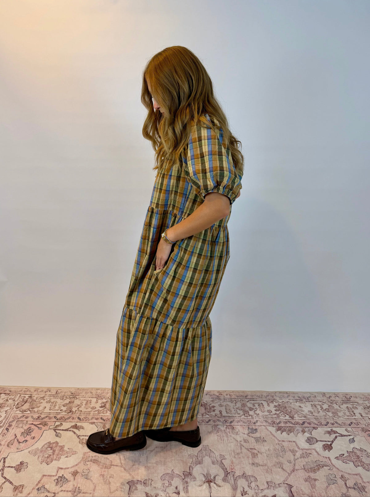 A woman wearing a long-sleeved plaid maxi dress with pockets, standing in profile against a plain white studio background.