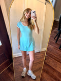 Person wearing a light blue romper taking a mirror selfie in a room with wooden flooring.