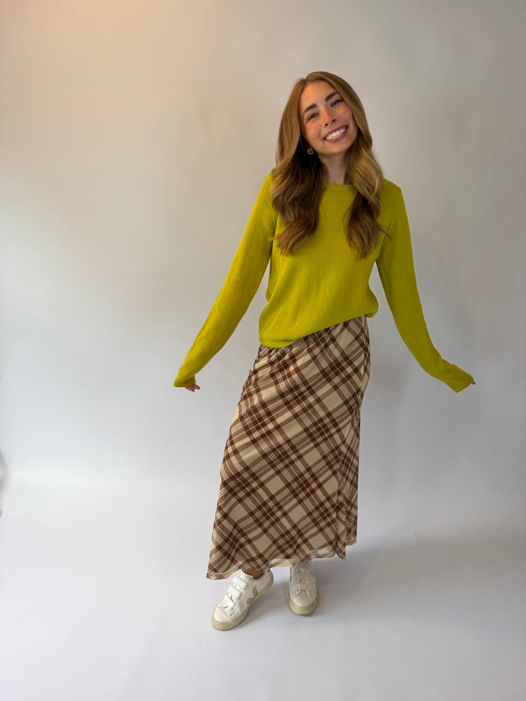 A woman modeling a bright yellow-green long-sleeve knit sweater and a brown and beige plaid maxi skirt, paired with white sneakers, against a plain light gray studio background.