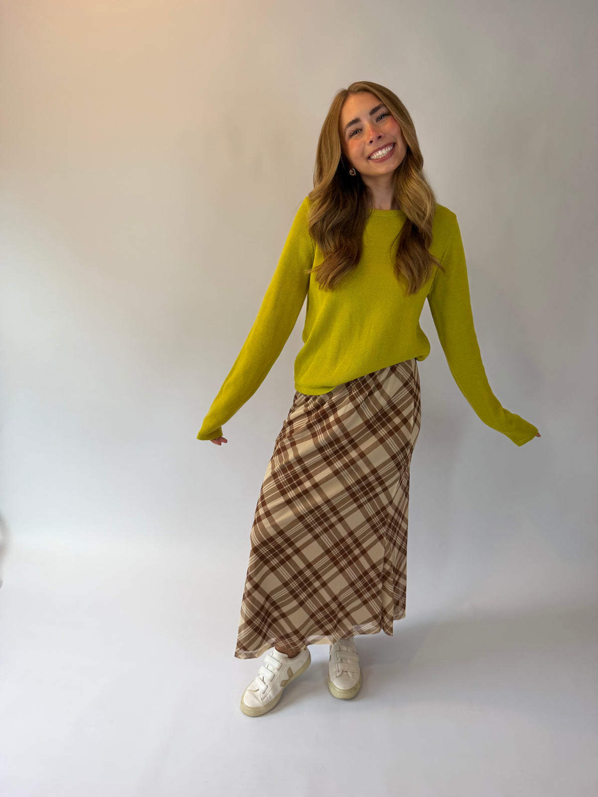 A woman modeling a bright yellow-green long-sleeve knit sweater and a brown and beige plaid maxi skirt, paired with white sneakers, against a plain light gray studio background.