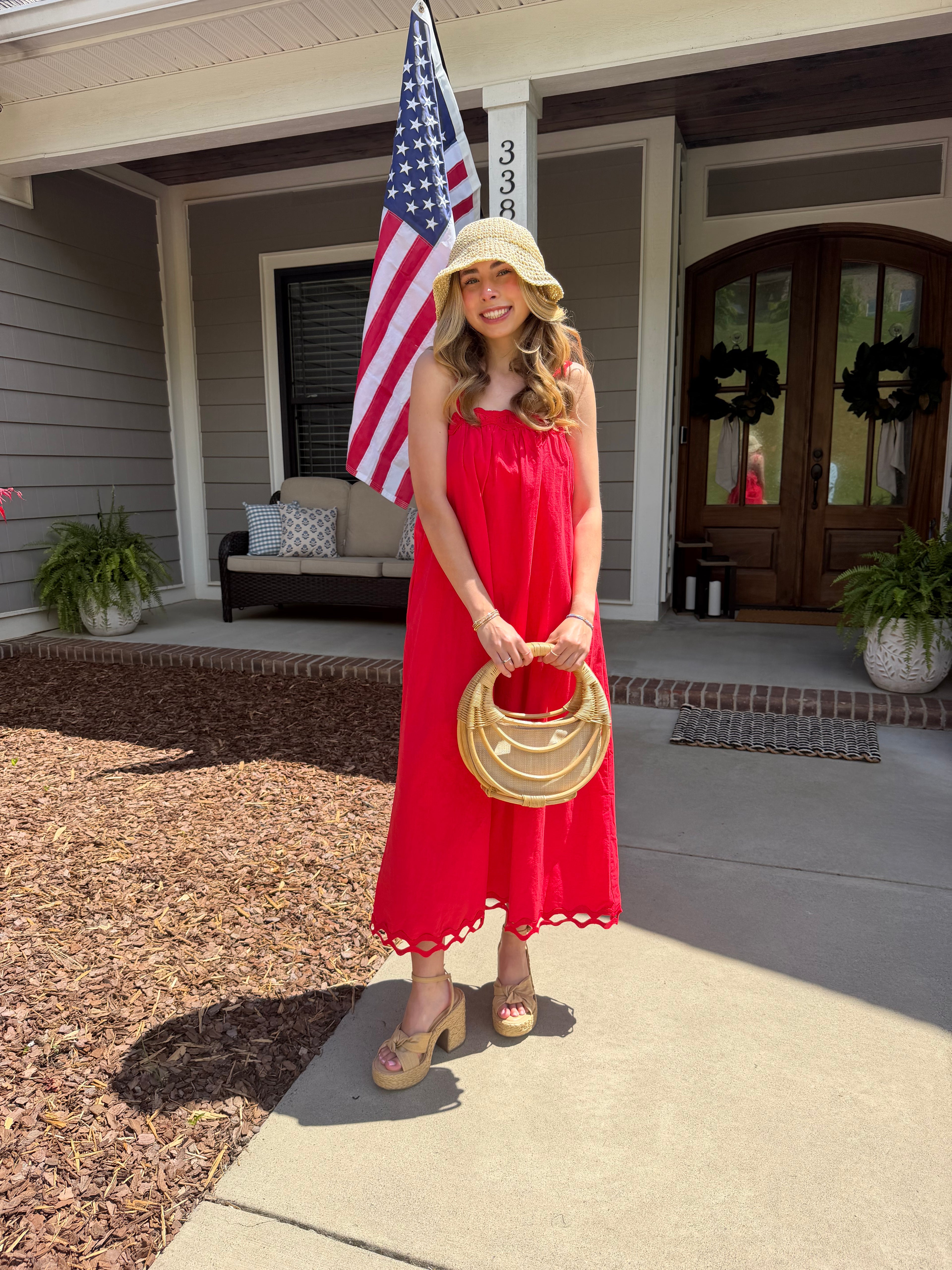 Woman in a red dress holding a gold handbag on a front porch with an American flag in the background.