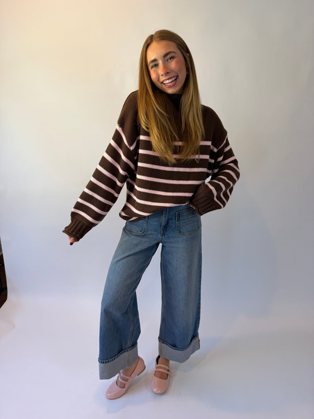 A woman wearing a brown and pink striped knit sweater, blue jeans, and pink ballet flats, photographed against a plain light background.