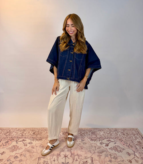 A woman modeling a dark indigo denim jacket with wide, short sleeves and cream wide-leg trousers, paired with white and black Mary Jane-style shoes, standing on a patterned rug against a neutral studio backdrop.