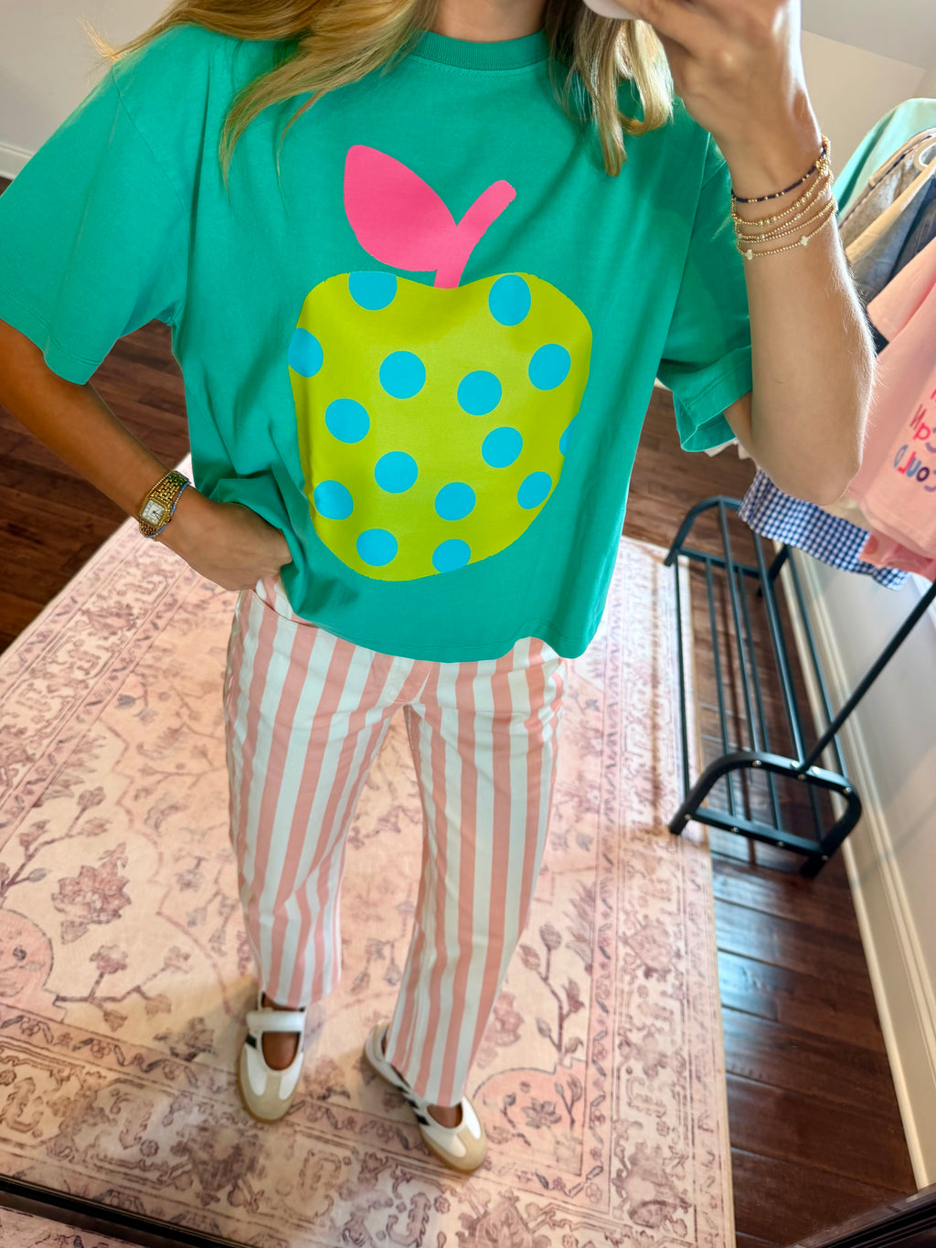 A person taking a mirror selfie in a room wearing a bright green graphic t-shirt with a large yellow-green polka dot apple design and pink leaf, paired with pink and white striped pants.