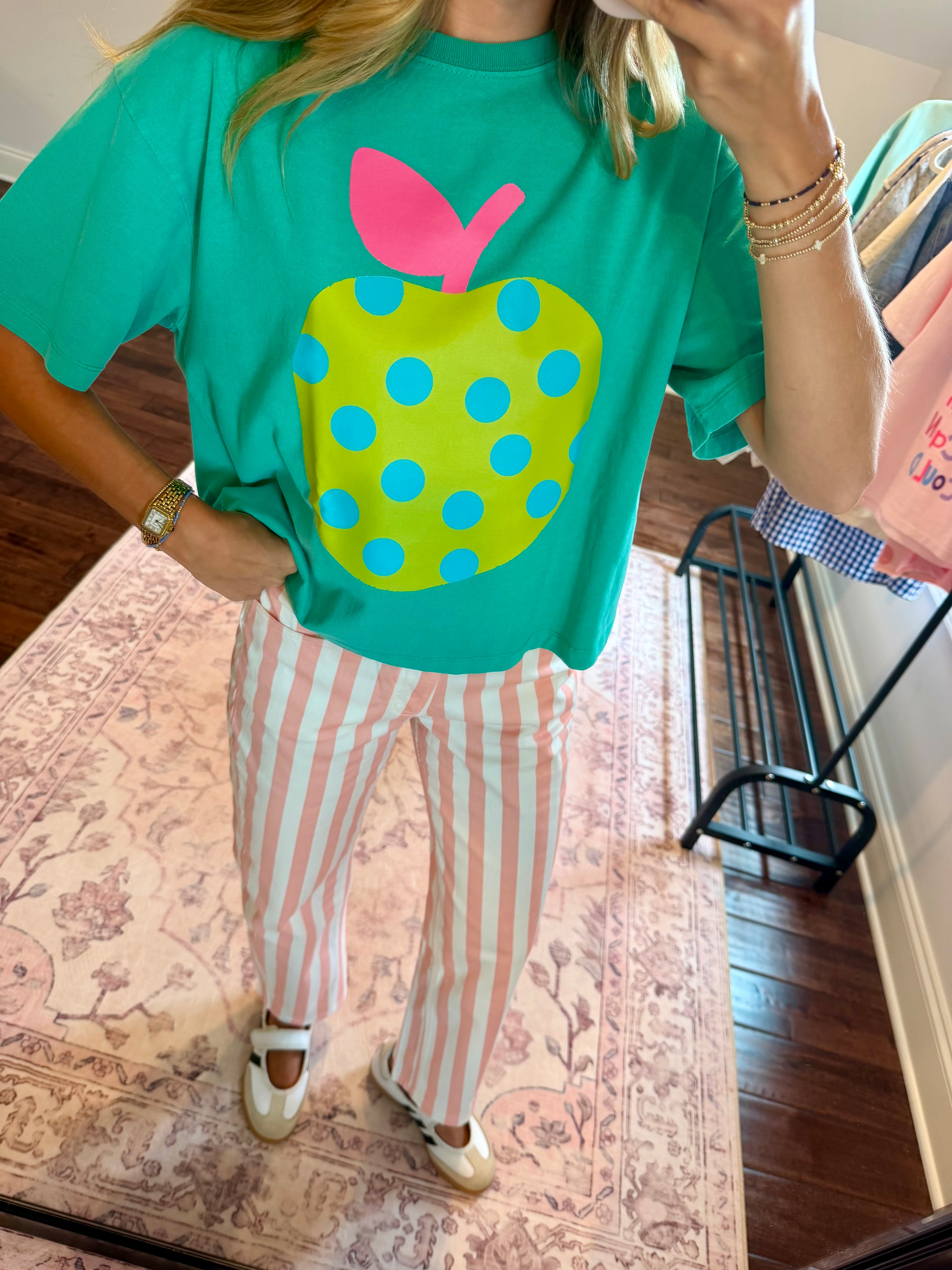 A person taking a mirror selfie in a room wearing a bright green graphic t-shirt with a large yellow-green polka dot apple design and pink leaf, paired with pink and white striped pants.