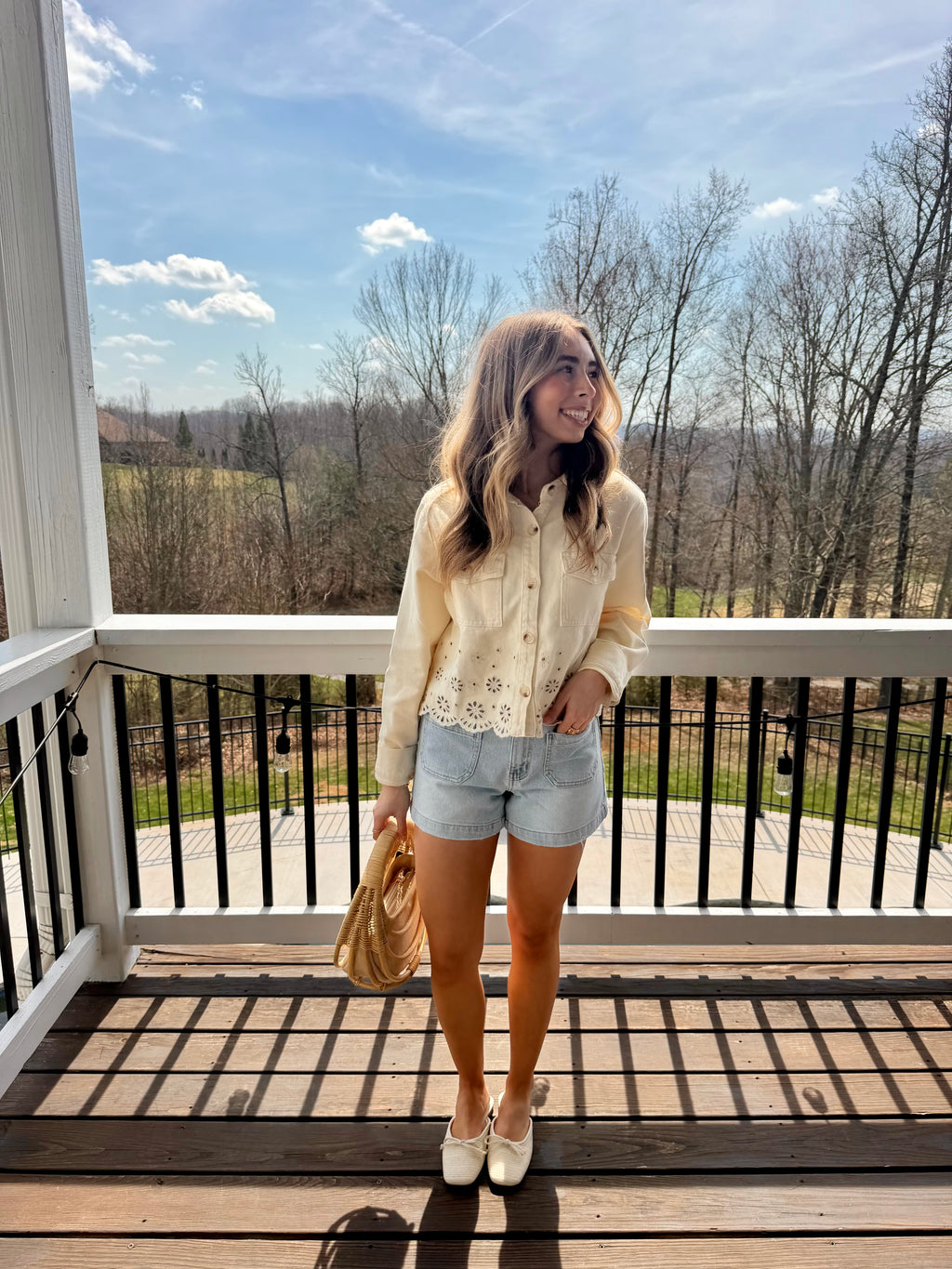 Woman standing on a deck with a scenic view, wearing a light-colored cardigan and shorts.