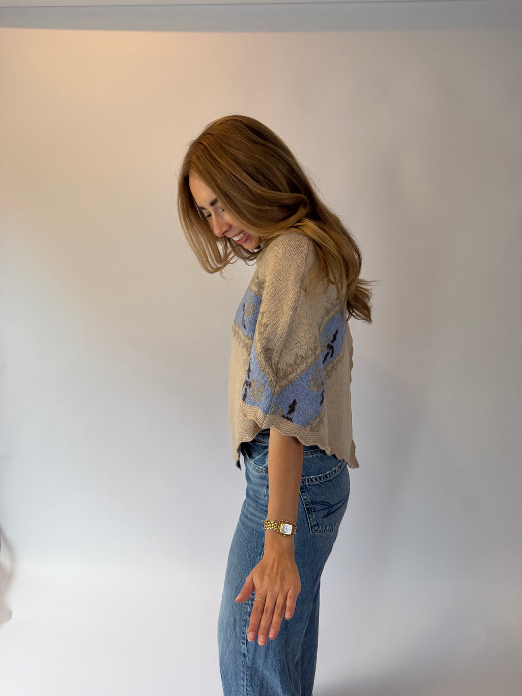 A woman with long light brown hair is shown from the side, wearing a beige knit top with blue and black abstract patterns on the sleeves, paired with blue jeans, against a plain white studio background.