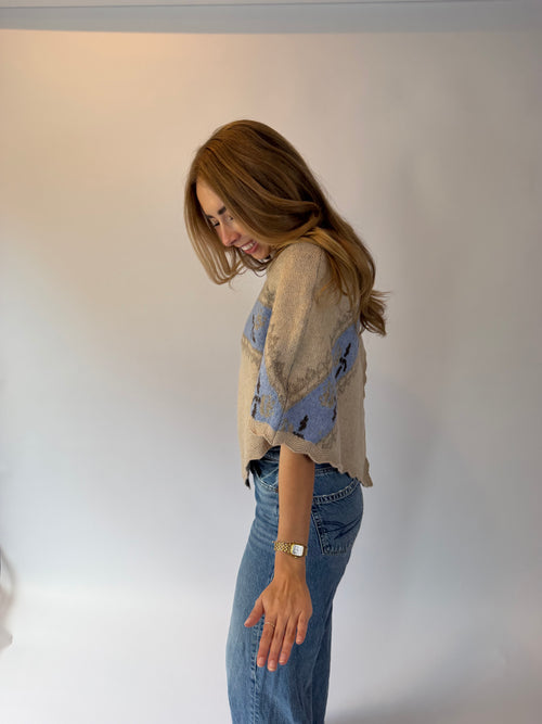 A woman with long light brown hair is shown from the side, wearing a beige knit top with blue and black abstract patterns on the sleeves, paired with blue jeans, against a plain white studio background.