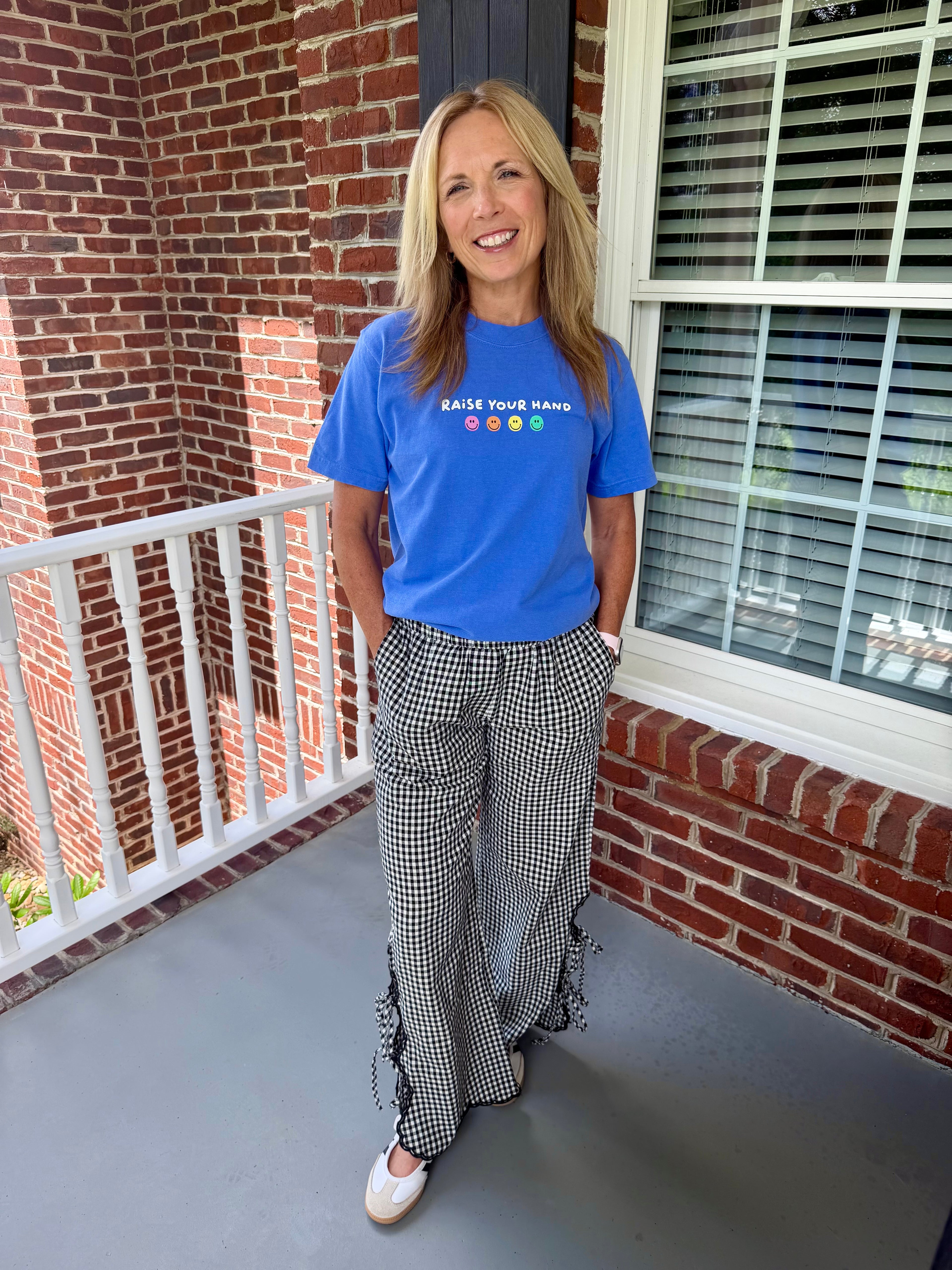 A woman stands on a home porch wearing a bright blue graphic t-shirt that reads 'RAISE YOUR HAND' with colorful hand silhouettes, paired with black and white gingham wide-leg pants with lace-up details at the hem and white sneakers.