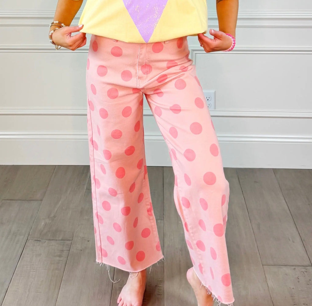 Pink wide-leg cropped pants with a pink polka dot pattern, shown on a model standing on a wood floor against a white paneled wall.