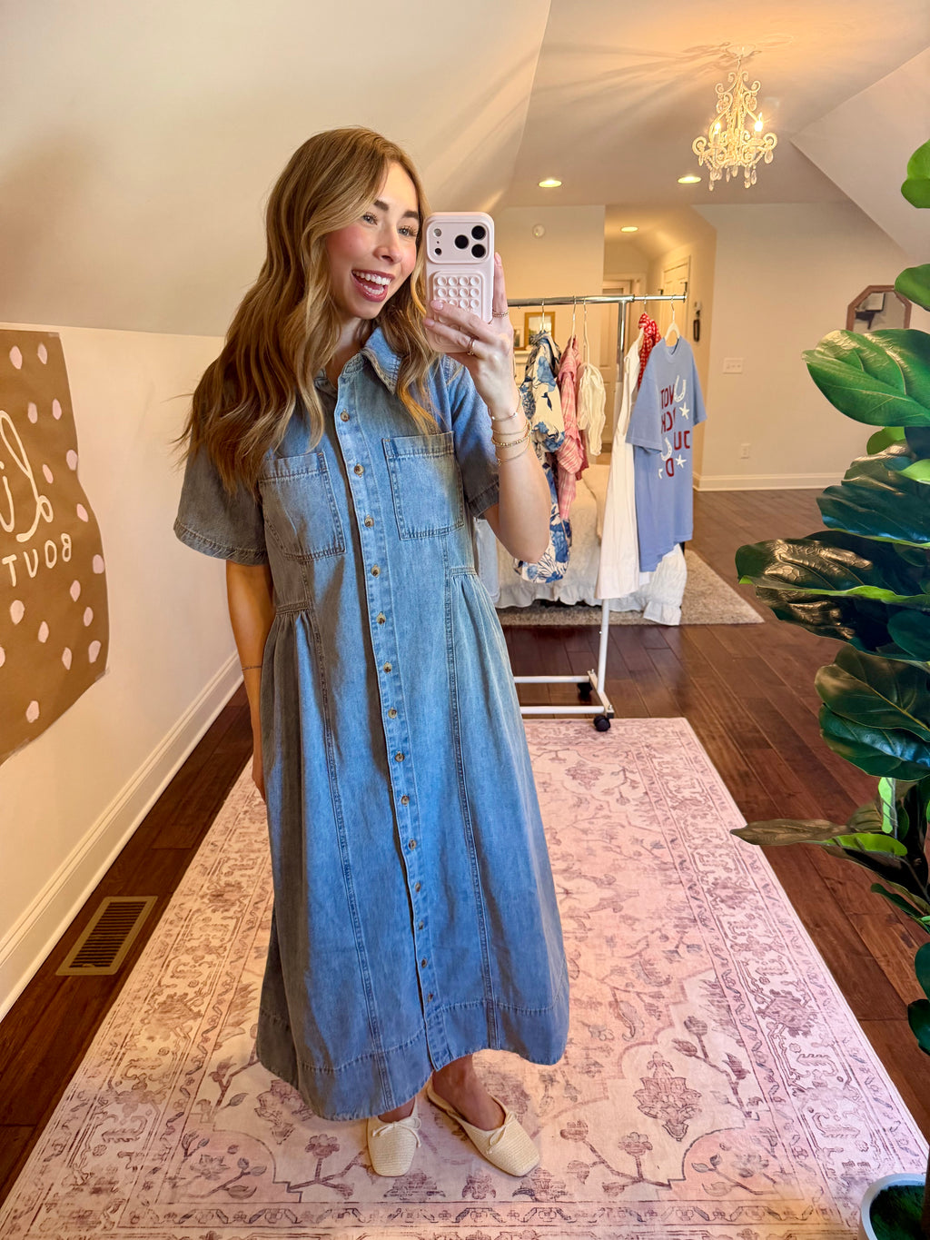 Woman taking a mirror selfie wearing a blue dress in a room with a chandelier and clothing rack.