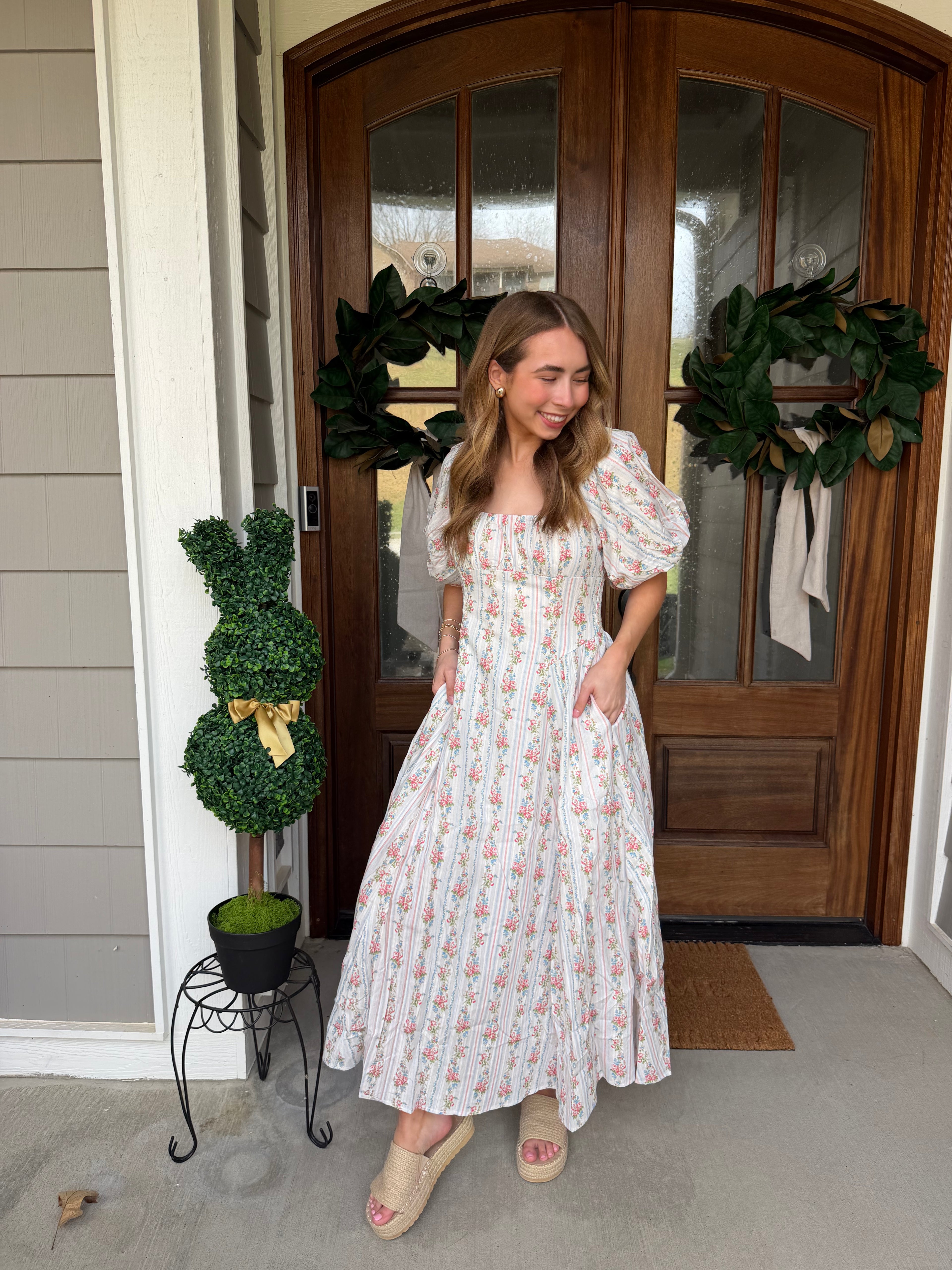 Woman in a white floral dress standing in front of a wooden door with wreaths.