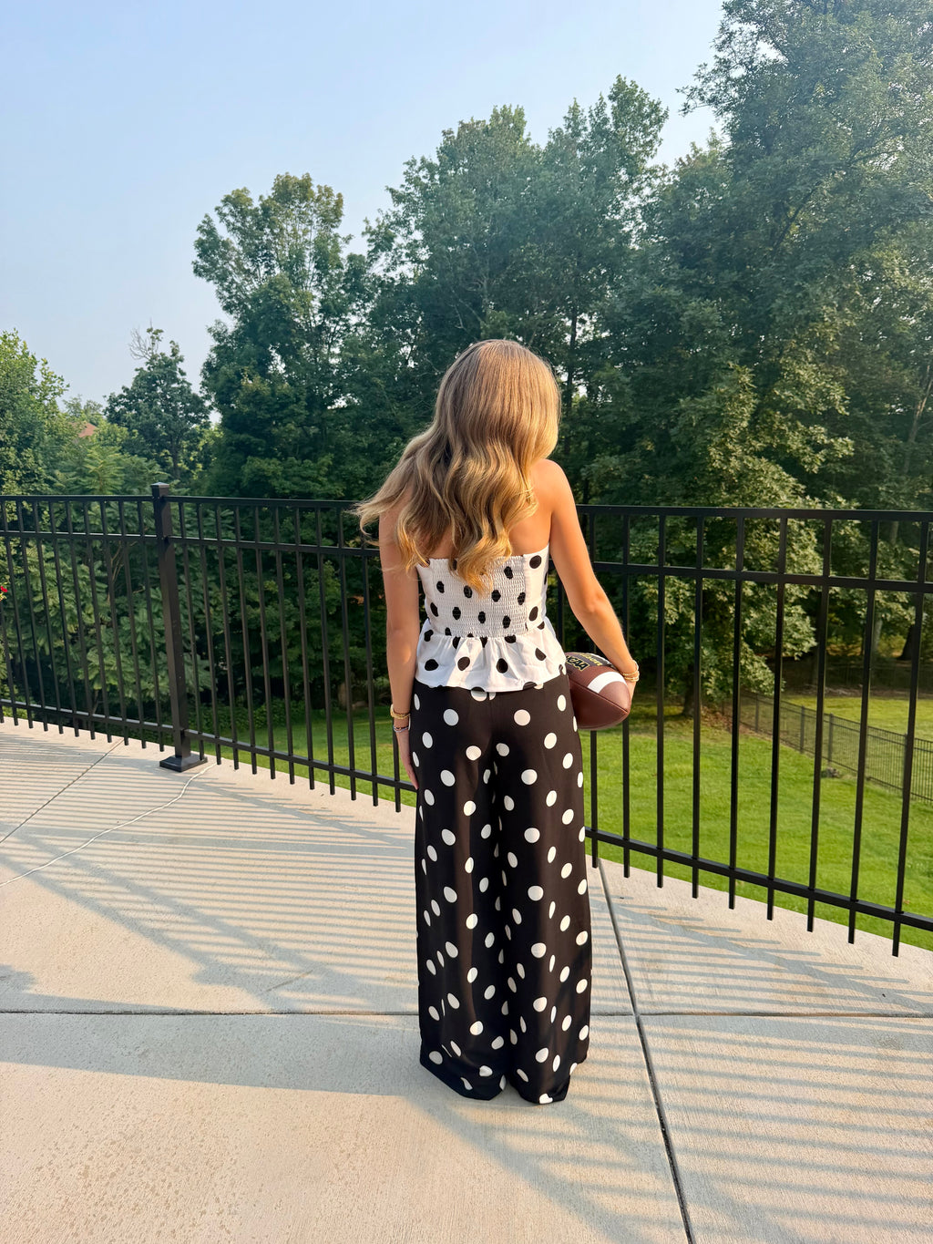 A woman stands on an outdoor patio holding a football, viewed from behind. She is wearing a white strapless top with black polka dots and a long black skirt with large white polka dots.