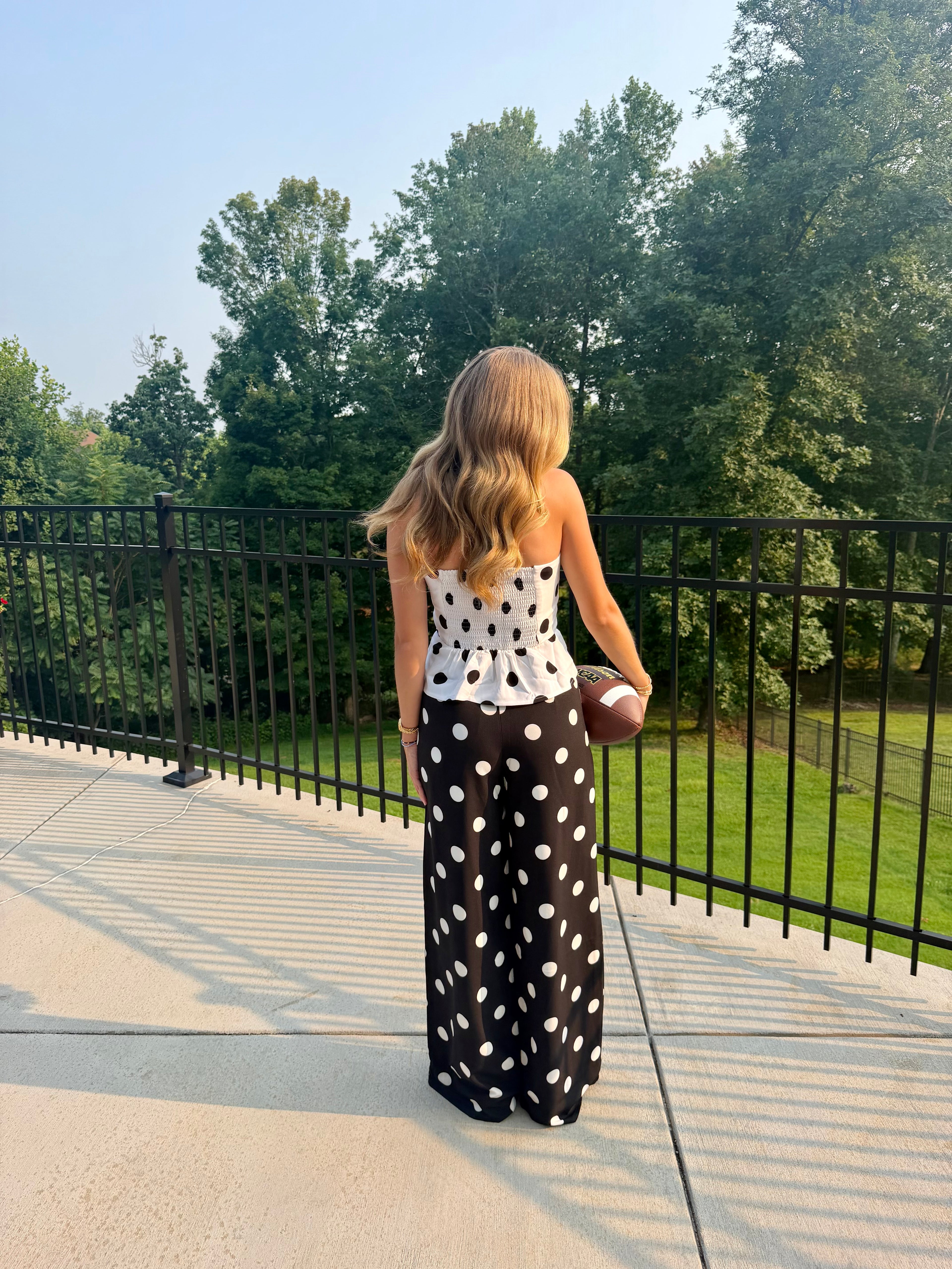 A woman stands on an outdoor patio holding a football, viewed from behind. She is wearing a white strapless top with black polka dots and a long black skirt with large white polka dots.