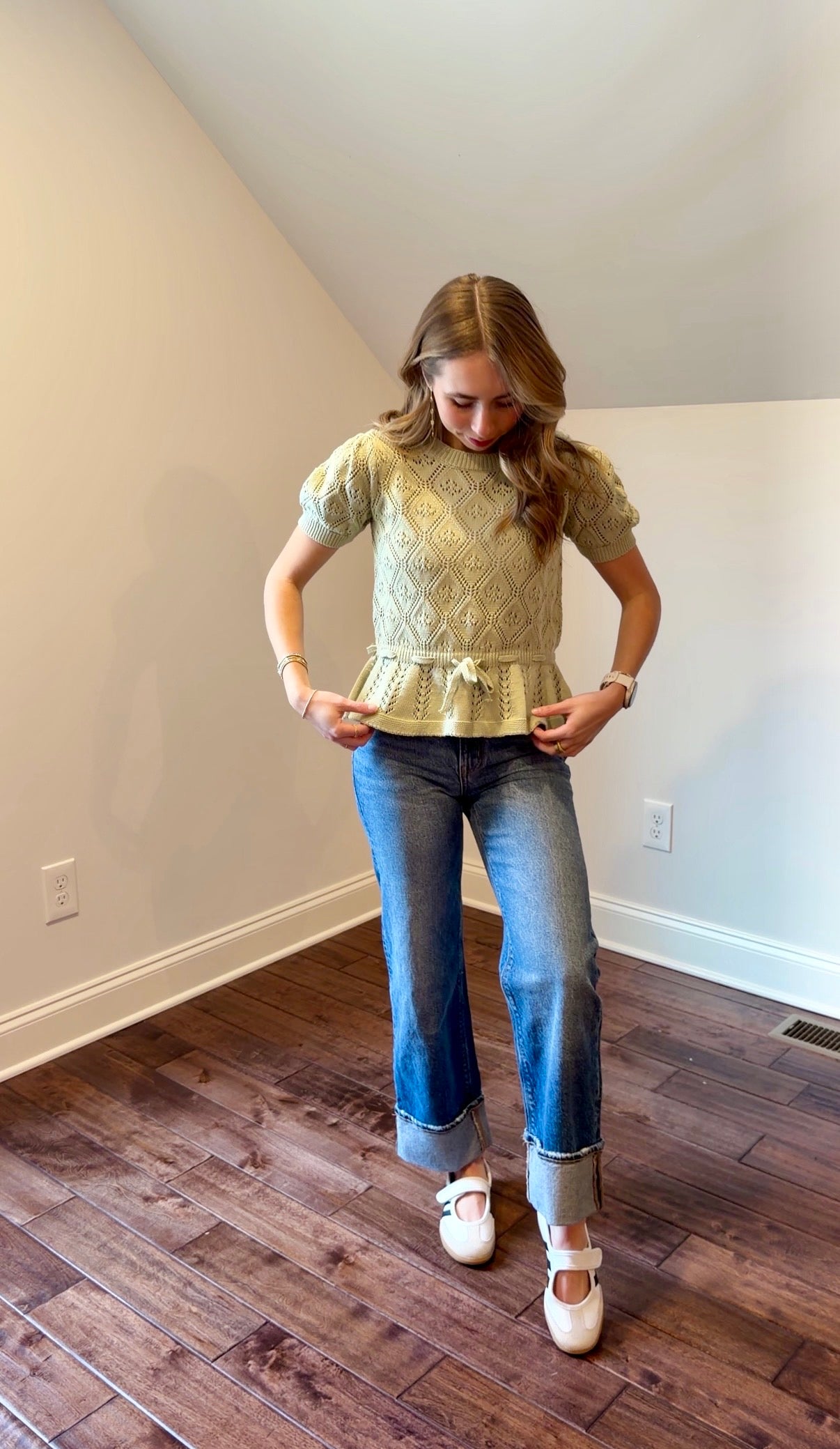 Woman wearing a green peplum top and blue jeans in a room with wooden flooring and white walls.